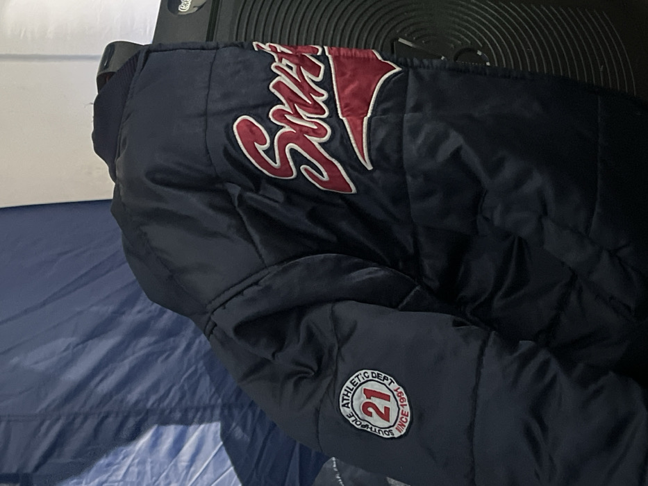 A dark blue quilted puffer jacket is depicted in an unknown, indoor or sheltered location within the city of Unknown, Unknown. The jacket is the primary subject, featuring a circular embroidered patch on its left sleeve displaying "SOUTHPOLE ATHLETIC DEPT" around the perimeter, a large red "21" in the center, and "SINCE 1991" below. On the right front of the jacket, large red and white embroidered lettering clearly spells "Southpole". The background on the left consists of a folded dark blue fabric, resembling a tent or tarp. To the right, a black, ribbed object, consistent with a speaker or fan unit, is partially visible, topped by a section of a white wall. No individuals, actions, or interactions are present in the image.