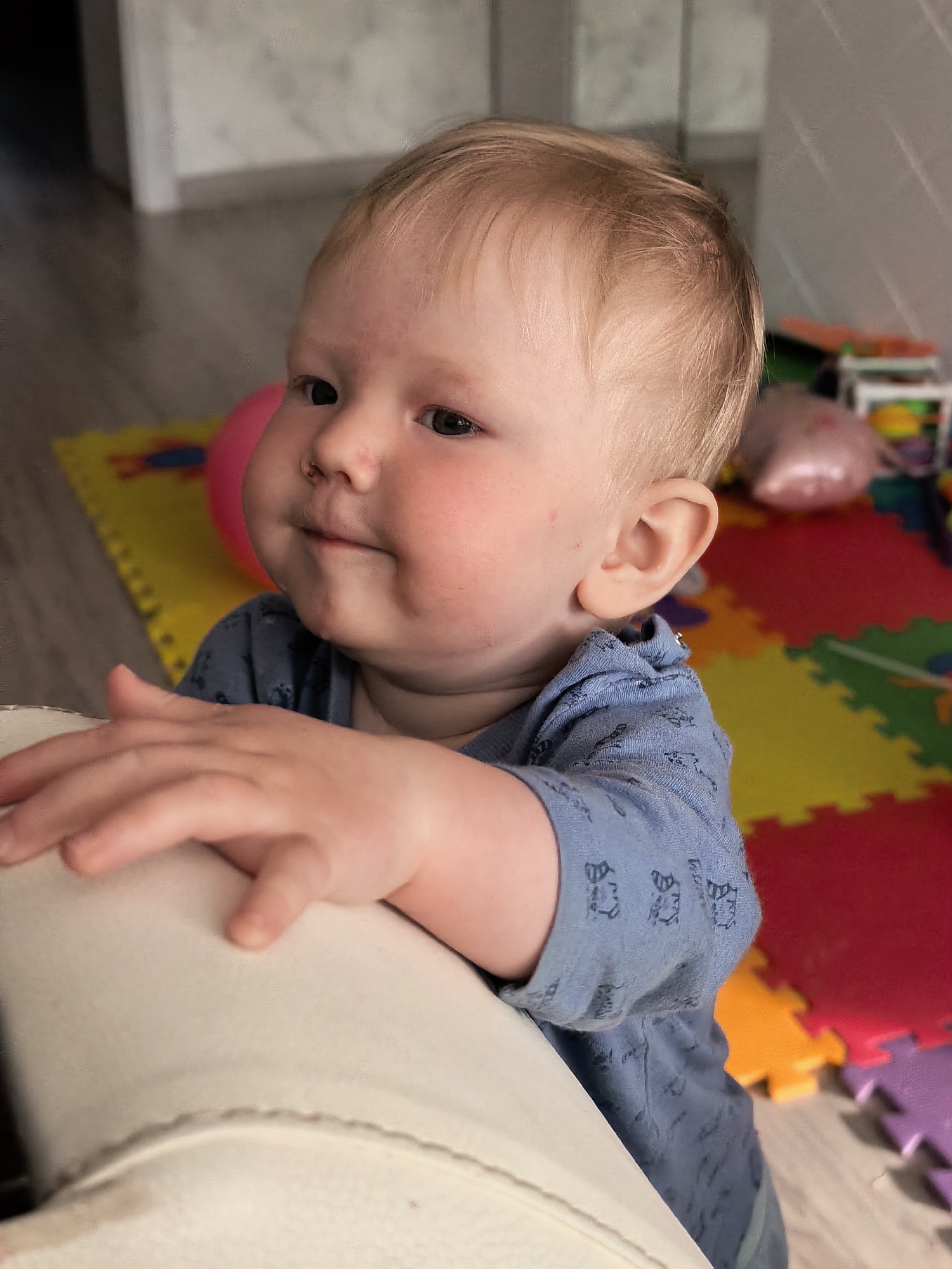 This is a medium close-up, vertical photograph featuring a fair-skinned baby with light blonde hair, likely a boy between 9 and 18 months old. The baby is positioned in the lower right portion of the frame, looking thoughtfully towards his left (viewer's right) with a slight, gentle smile or smirk. His full cheeks are rosy, and a few small, faint reddish marks are visible on his face, including his forehead, cheeks, and a mark on his nose.
He is wearing a long-sleeved blue shirt patterned with small, dark, repetitive designs, possibly cartoon animals or abstract shapes. The baby is standing and holding onto a light-colored (cream or white) piece of furniture, resembling the armrest of a sofa or chair, with his right hand prominent in the foreground.
The background depicts a vibrant indoor play area. The floor is covered with colorful interlocking foam play mats in red, yellow, green, orange, and purple. A bright pink inflatable ball is visible behind the baby's head, and other blurred toys and household items are scattered further back. The overall lighting suggests it is daytime in a residential setting, likely a home in Sterlitamak, Russia. There is no visible text in the image.