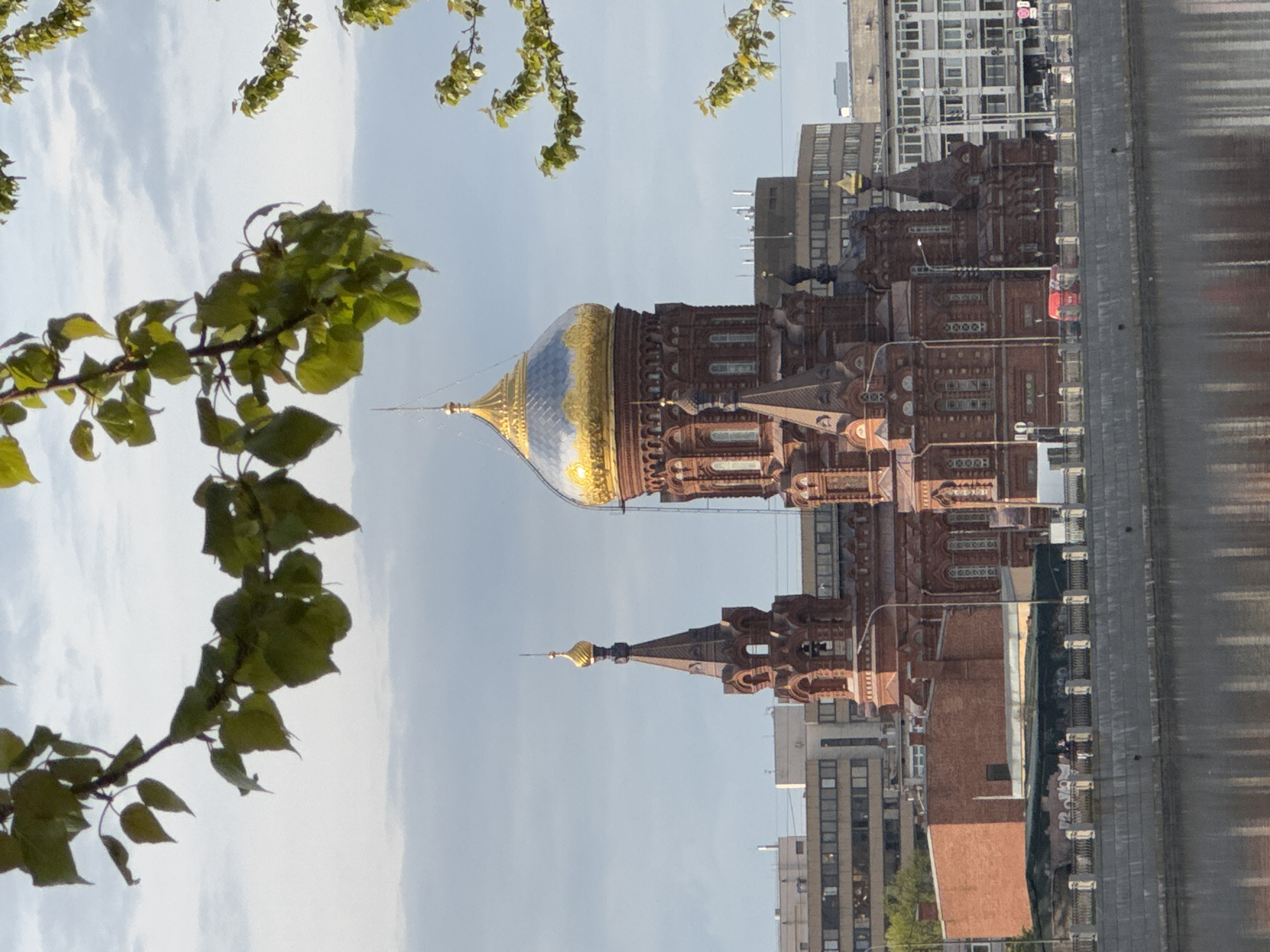 The image depicts a view of the Church of the Savior on Spilled Blood in Saint Petersburg, Russia, seen through foliage. The architectural details of the church are visible, including its ornate brickwork, domes, and spires. The larger dome is notable for its gold and blue coloration. The church is situated in an urban environment with surrounding buildings, and a body of water is visible in the foreground, potentially a canal.