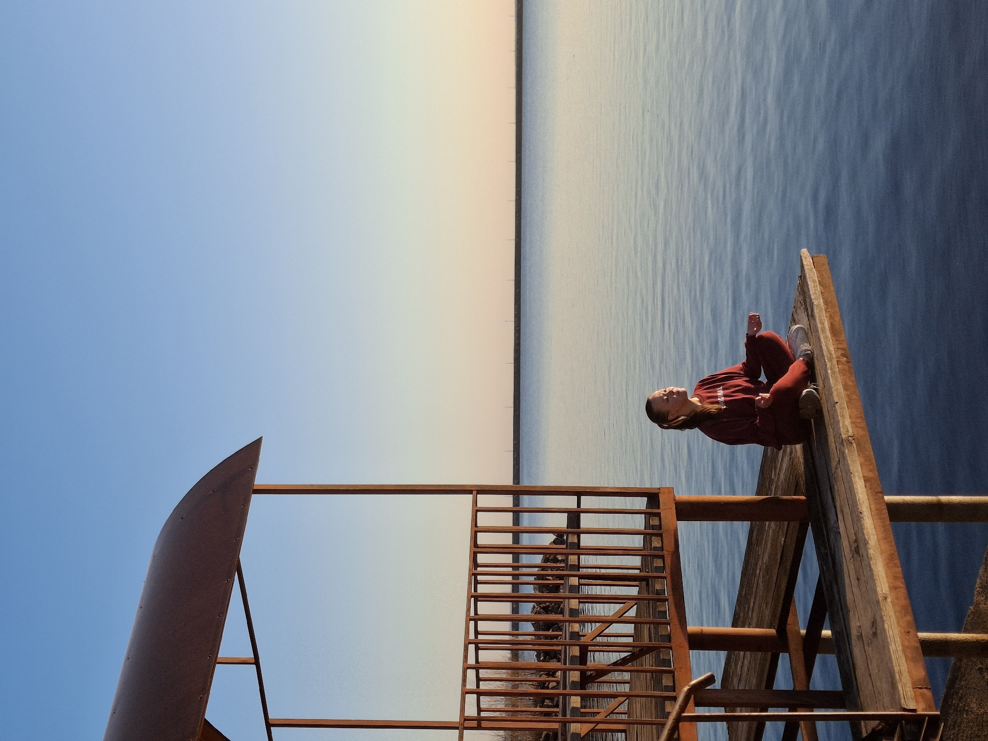 A woman is sitting on a wooden structure next to a body of water, possibly a lake or a wide river, in Chaikovsky, Russia. She is wearing a reddish-brown sweat suit and sneakers. The structure she is sitting on appears to be part of a rusted metal and wooden pier or platform. The woman seems to be looking out at the water, with her head tilted upwards. The sky is clear with a gradient of blue to a light peach color near the horizon. In the distance, there is a dark line, possibly a coastline or the opposite bank of the water body. The architectural elements of the pier or platform include rusted metal supports, a staircase-like structure, and a curved piece of metal.