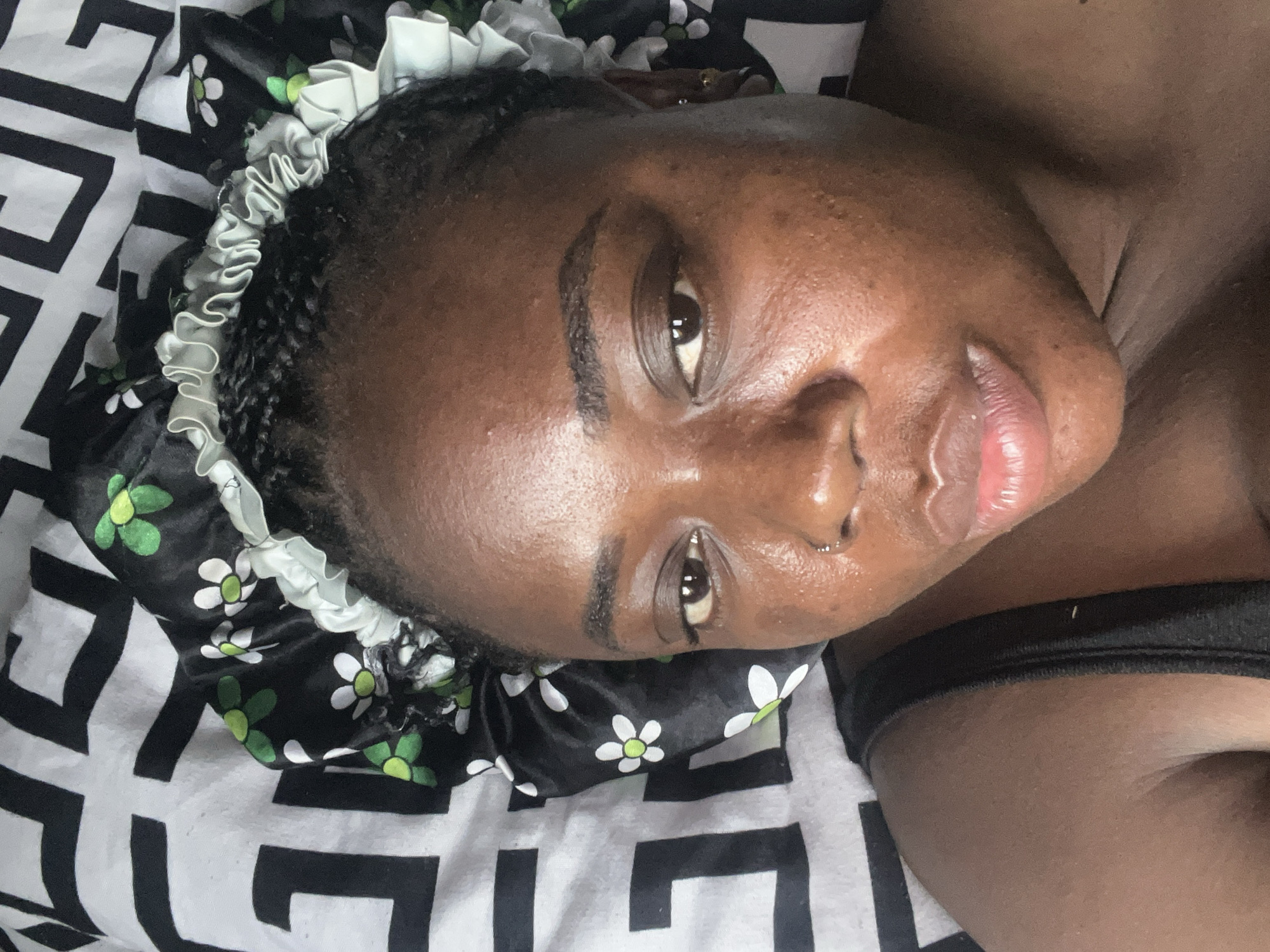A close-up, slightly rotated, high-angle shot of a Black woman with dark skin, lying down. She is wearing a black and white patterned satin bonnet with green and white flower details and a white ruffled edge. The pattern of the bedsheet she is lying on has a bold black and white geometric design. Her hair is braided underneath the bonnet. Her facial features are prominent, including her dark eyes, nose with a visible piercing on the left side, and slightly parted lips with a glossy appearance. A thin black strap of a garment is visible near her shoulder. The location and city of Abuja, Nigeria, are not visually evident.