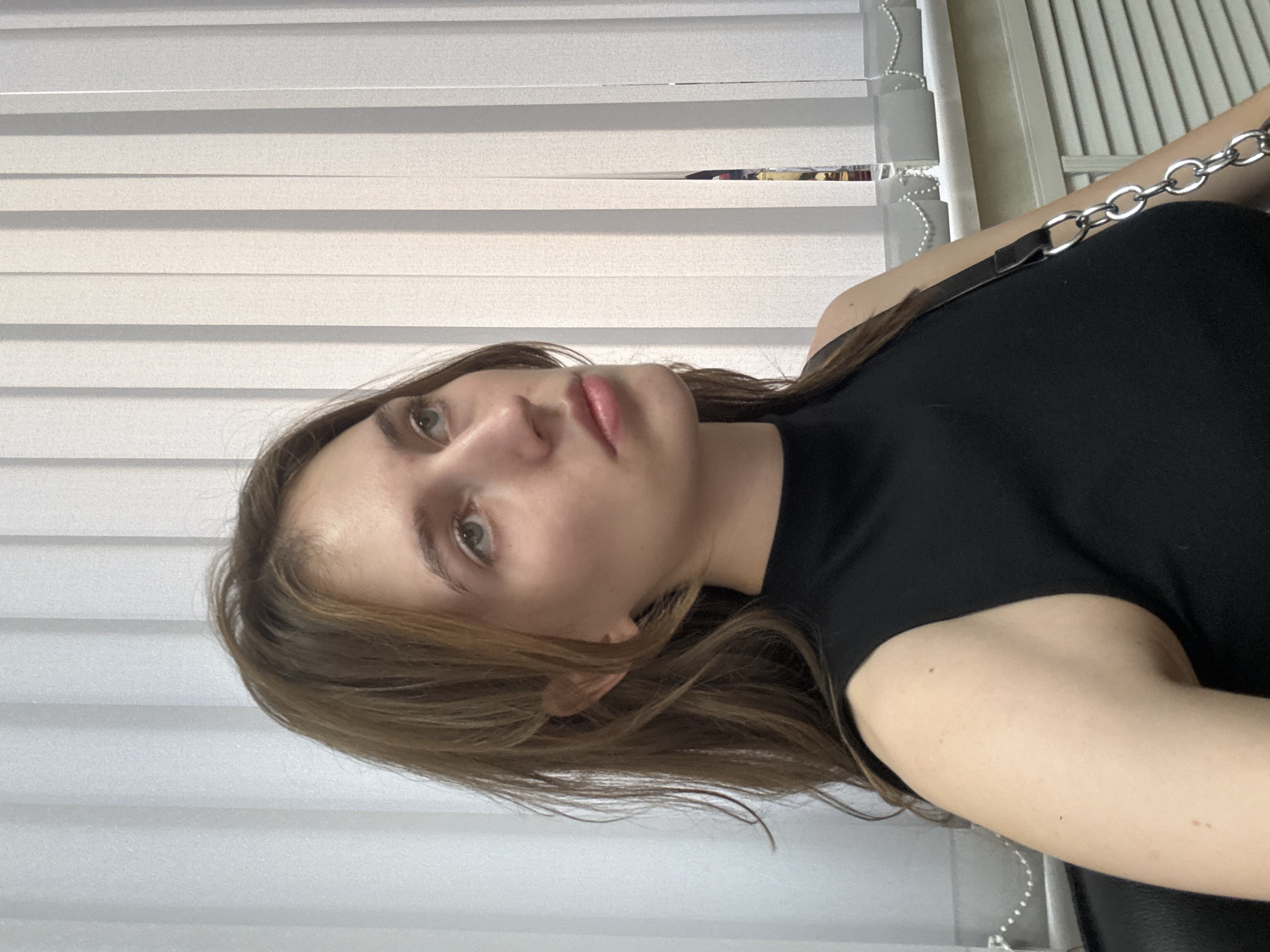 A young woman with shoulder-length brown hair is captured in a medium-close shot, looking upwards and to her right. She is wearing a black sleeveless top and a black purse with a silver chain strap is visible. The background consists of vertical white blinds, typical of an indoor setting, possibly an office or apartment. The lighting suggests an indoor environment with natural light filtering through the blinds. The exact location in Perm, Russia, cannot be determined from the visual information provided.
