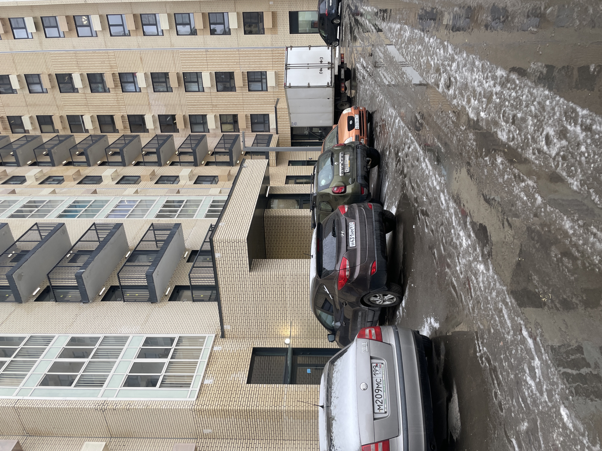 The image shows a street scene in Unknown, Unknown. The street is wet, and there is some ice or slush on the ground. A tall brick building lines the left side of the street. Several cars are parked along the right side of the street. They are a silver car, a brown SUV, a green SUV, an orange car, and a white truck. A black car is parked in the distance.