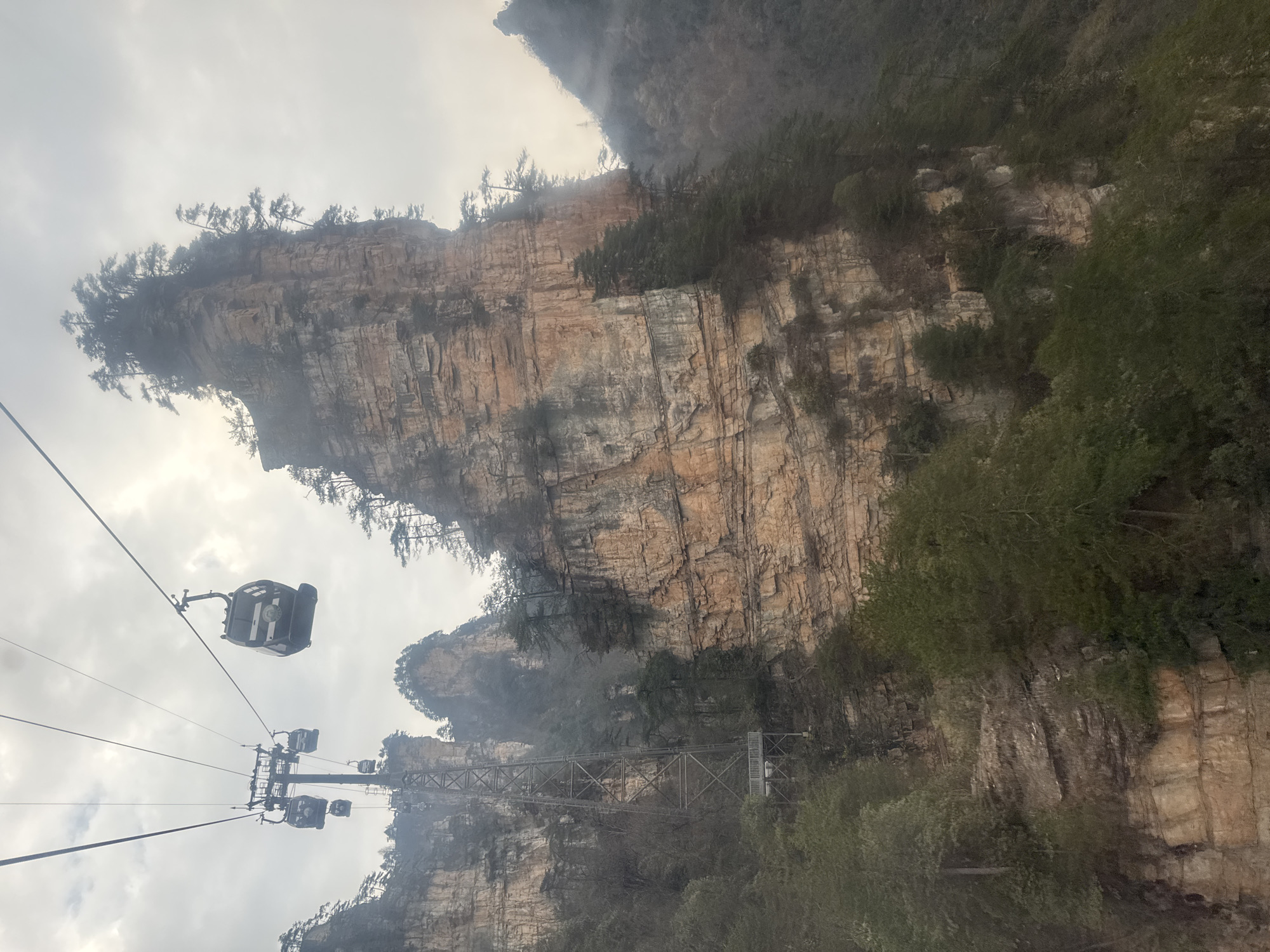 This image depicts a gondola lift system traversing a rugged, mountainous landscape in Wulingyuan, China. The scene features multiple dark-colored gondola cabins suspended from parallel cables under an overcast, hazy sky. One gondola cabin, with distinctive white or light gray horizontal striping, is prominent in the upper right. A tall, gray, lattice-structured metal support tower for the gondola system stands prominently in the mid-ground on the left, anchored to a rocky slope and featuring a small platform at its base.
The environment is characterized by towering, sheer sandstone or quartz-sandstone pillars and cliffs, displaying clear horizontal stratification in shades of beige, brown, and gray. These precipitous rock faces are sparsely covered with dark green evergreen trees, primarily pines, growing from crevices and atop the peaks. Dense foliage and additional rocky terrain are visible below the gondola lines. The atmospheric conditions include diffused light and pervasive fog or low-hanging clouds, partially obscuring distant peaks and creating a muted palette. No individuals are discernible within the gondola cabins or anywhere else in the scene.