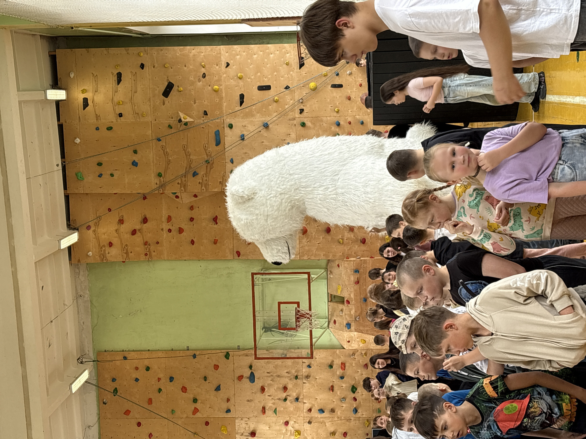 The image appears to be taken inside a school gymnasium or sports hall in Izhevsk, Russia. The dominant feature is a climbing wall with numerous colored handholds. A large, white polar bear mascot head is present among a crowd of children and adults. The people are diverse in age, with students and possibly teachers or guardians present. A basketball hoop is visible on the wall. The group seems to be engaged in an event or assembly of some kind.