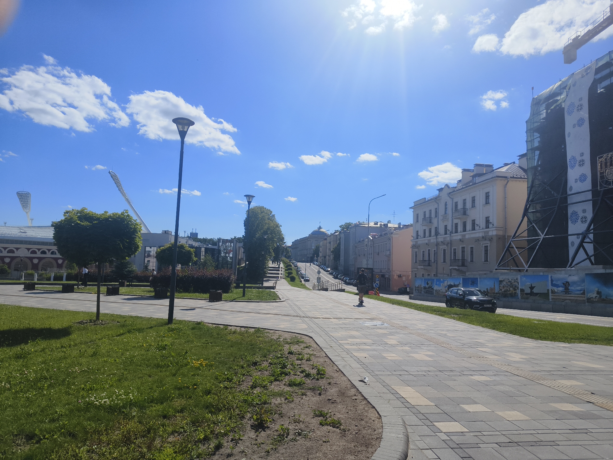 The image depicts a sunny day on a wide, paved pedestrian walkway in Minsk, Belarus. The walkway, laid with light and dark gray rectangular pavers, curves to the right. On the left, a grassy area slopes down, with a lamppost and manicured trees. In the background, a large building with arched windows and a distinctive stadium-like structure with floodlights is visible.
To the right of the walkway, a street runs parallel, with parked cars and a building under construction, covered in scaffolding and a large banner featuring blue snowflake-like designs and a coat of arms. Further down the street, a row of traditional European-style buildings with multiple stories and balconies lines the road.
A lone individual is rollerblading down the walkway. The sky is a vibrant blue with scattered white clouds, and the sun is shining brightly, casting lens flares and highlights across the scene. The overall impression is of an urban public space on a pleasant day.