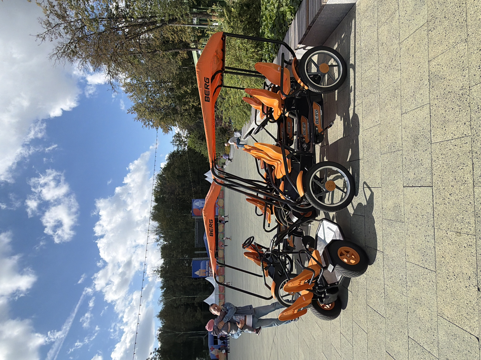 The image shows a sunny outdoor scene in a park-like setting, possibly a recreational area in Nizhny Novgorod, Russia. In the foreground, there are two orange pedal go-karts, each with a black frame and a sun canopy labeled "BERG." The go-karts have multiple seats and steering wheels. To the left of the go-karts, a man is holding a young child, who is wearing a pink hat. The man is dressed in a denim jacket and jeans. Behind the man and the go-karts, there is a paved area with some distant figures and tents. Further in the background, there are trees with green and some autumn-colored foliage, a blue sky with white clouds, and what appears to be a stage or event setup with banners. The overall atmosphere suggests a public event or a place where families can rent and enjoy outdoor activities.