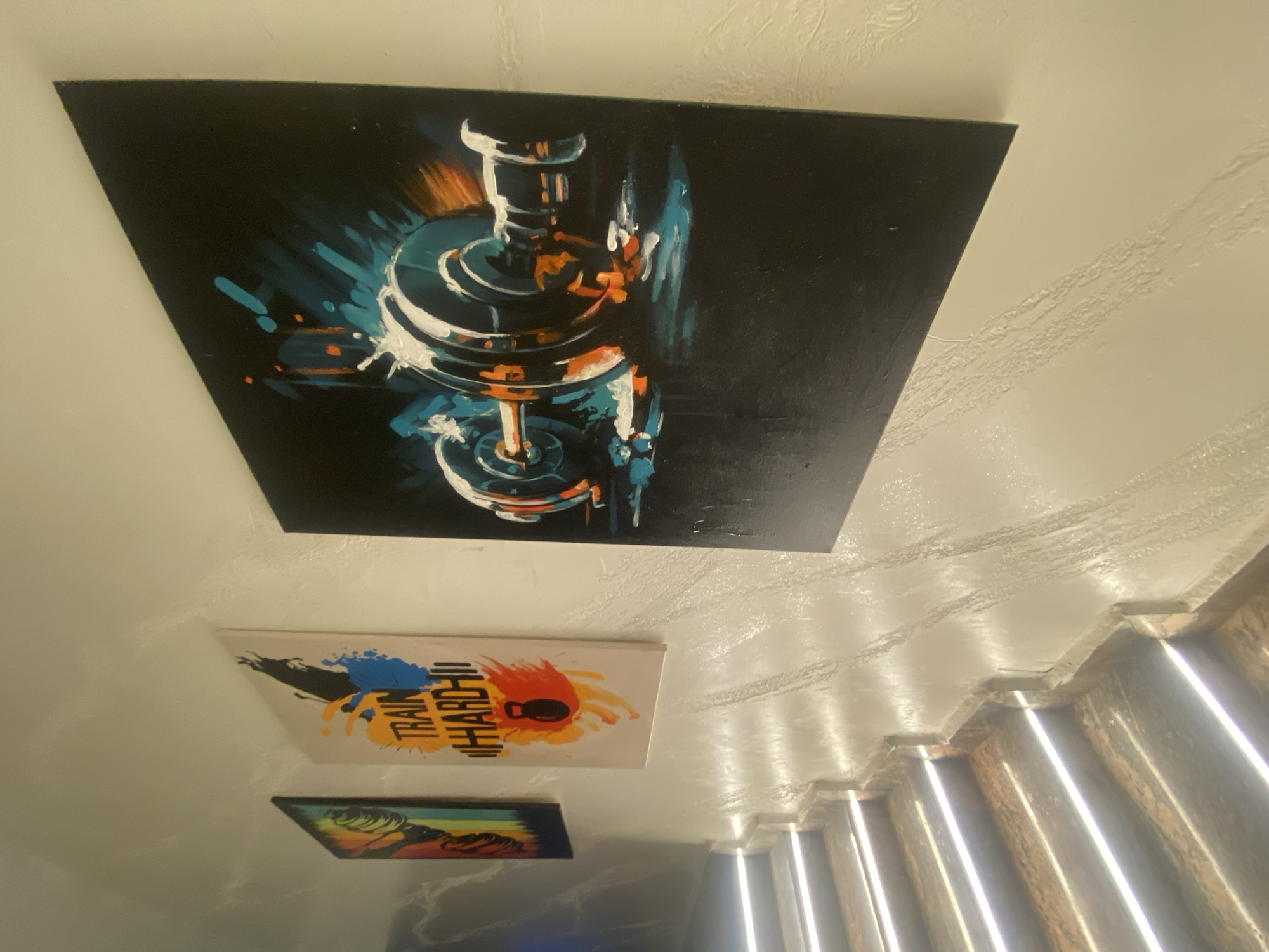 The image displays a brightly lit staircase with modern, integrated LED lighting. The walls adjacent to the staircase are textured and painted in a light color. Several pieces of framed artwork are hung on the wall. The artwork consists of vibrant, abstract depictions of fitness-related themes. One prominent painting features a dumbbell with dynamic, energetic brushstrokes of blue, orange, and white against a black background. Another painting displays the text "TRAIN HARD" with an illustration of a kettlebell, rendered in bold yellow, orange, and blue splashes. A third, partially visible artwork to the far left appears to be a colorful abstract piece. The scene suggests a contemporary interior design, possibly a gym, studio, or a home with a focus on fitness motivation. No individuals are present in the frame. The location is not specified as Barden Yana, Nigeria.