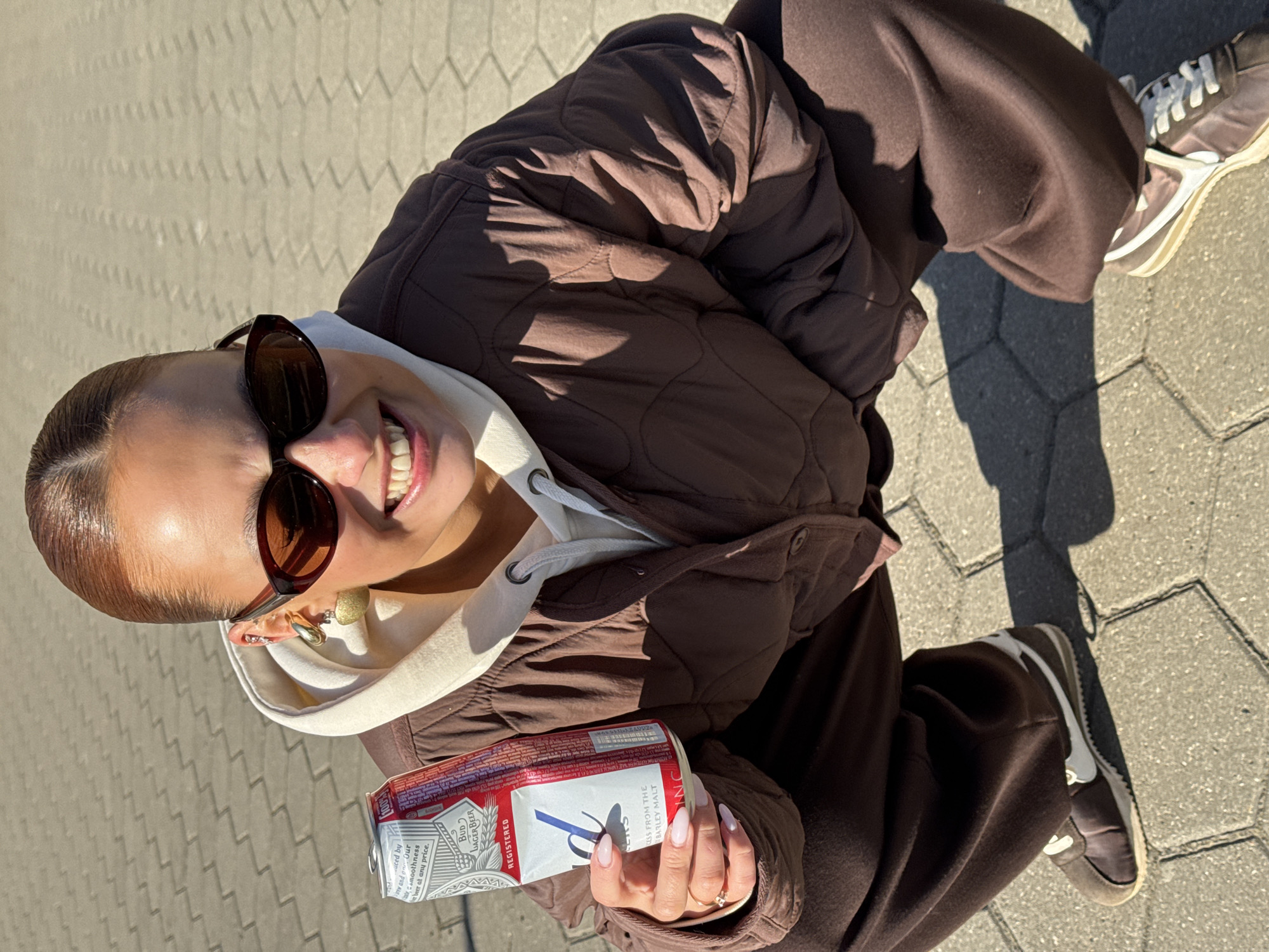 A woman is seen from an overhead angle in what appears to be an urban setting. She is smiling and wearing dark sunglasses, a light-colored hoodie beneath a brown, quilted jacket, and matching brown pants. Her nails are neatly manicured. She is holding a twelve-pack of Budweiser beer. The setting is paved with interlocking hexagonal blocks. Part of a brown and white sneaker is visible on the right side of the frame. The OCR text suggests the beer is promoted for its smoothness at any price, and that it contains "kiss from the barley malt." The location is not discernable from the image.