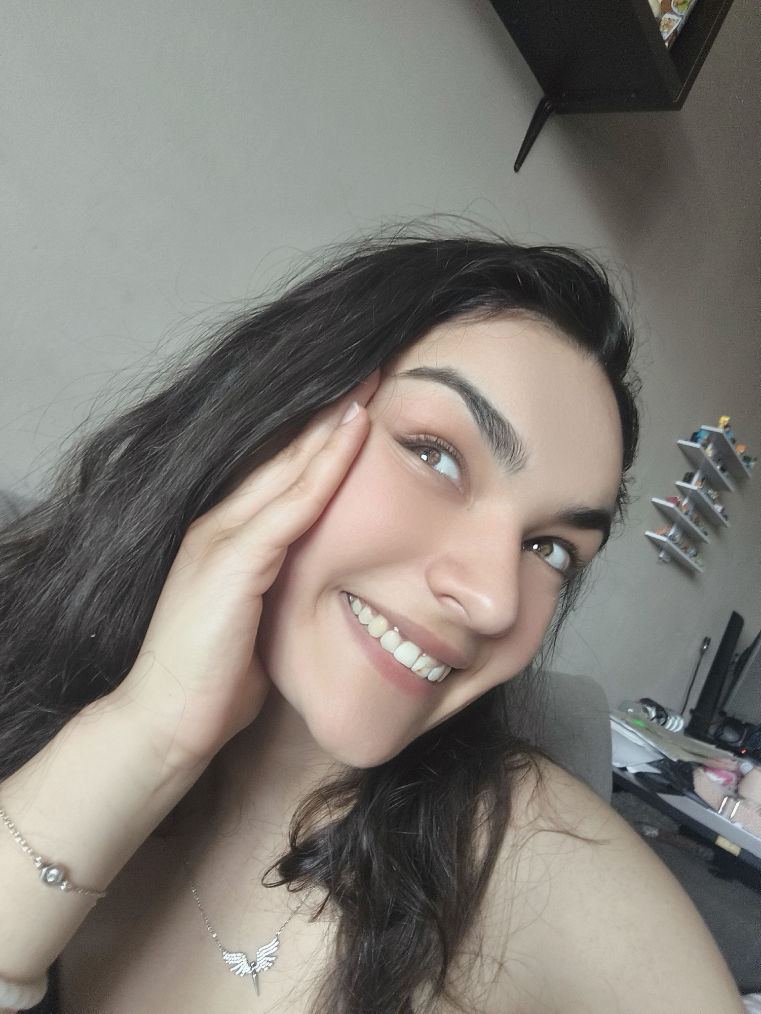 A young woman with long, dark hair and brown eyes is shown in a close-up selfie. She is smiling and her right hand is touching her face. She is wearing a silver necklace with a winged pendant and a bracelet. The background appears to be an indoor setting, possibly a living room, with a gray wall and a shelf with items on it. There is no indication of the city of Sofia, Bulgaria, in this image.