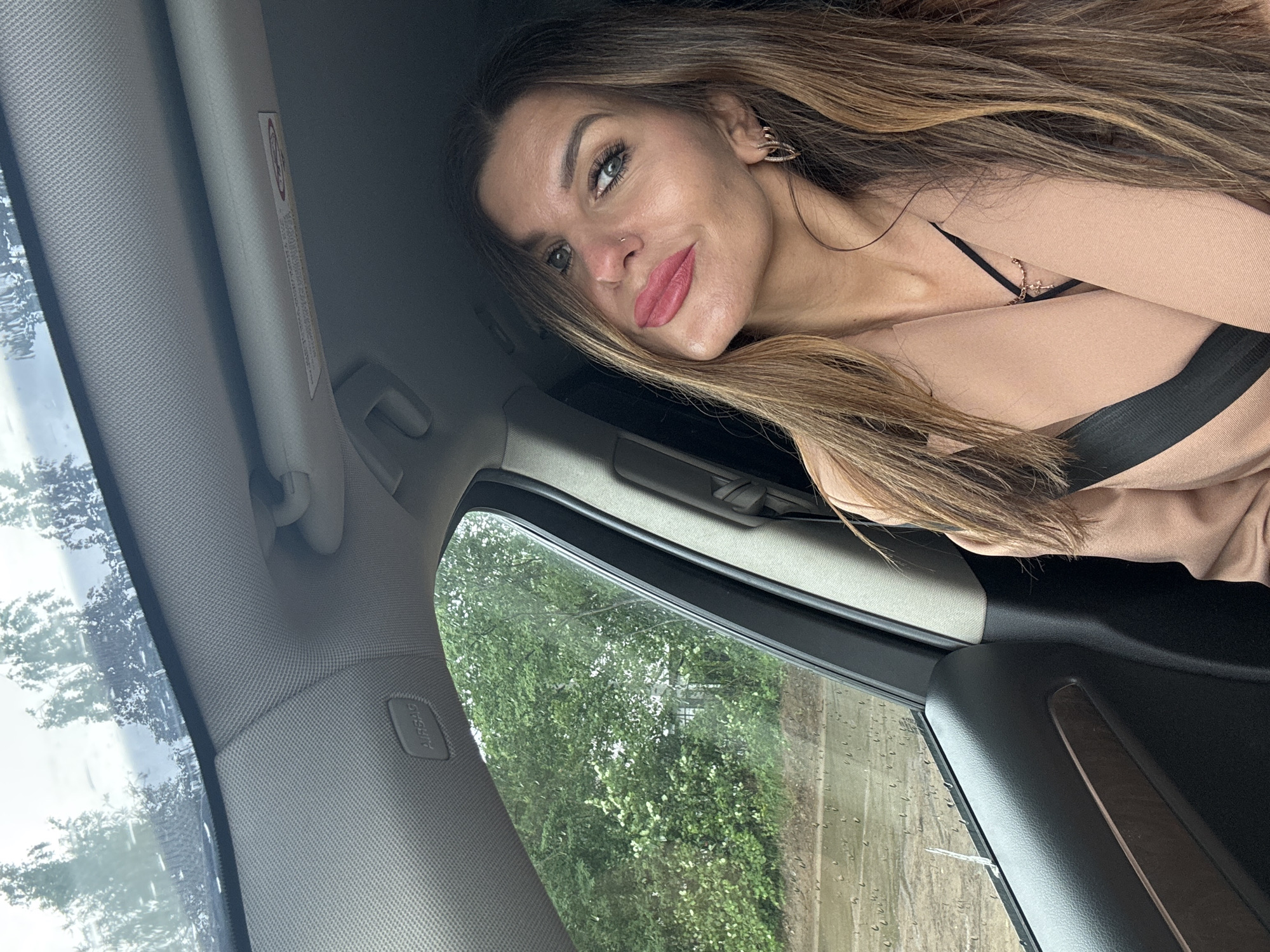 This medium shot captures a woman inside a car in Chaikovsky, Russia, posing for a photo. The image is taken at an angle, with the woman's head tilted, showing her from the chest up. She has long, light brown hair and is wearing a light beige or nude-colored top with a black strap visible. Her makeup includes defined eyebrows, dark eyeliner, and bright pink lipstick. She has a small nose piercing and a distinctive earring in her right ear, and her expression is a soft, direct gaze at the camera.
The setting is the interior of a vehicle, identifiable by the light grey textured headliner, a-pillar, and sun visor. An "AIRBAG" label is prominently visible on the a-pillar cover. A warning label on the sun visor shows a red circular symbol with a diagonal line, though the specific text is unreadable due to the angle and focus. Through the car windows, green trees and foliage are seen, indicating an outdoor, natural environment. Raindrops on the side window suggest that it is either raining or has recently rained. The time of day is clearly daylight, with bright natural light illuminating the scene.