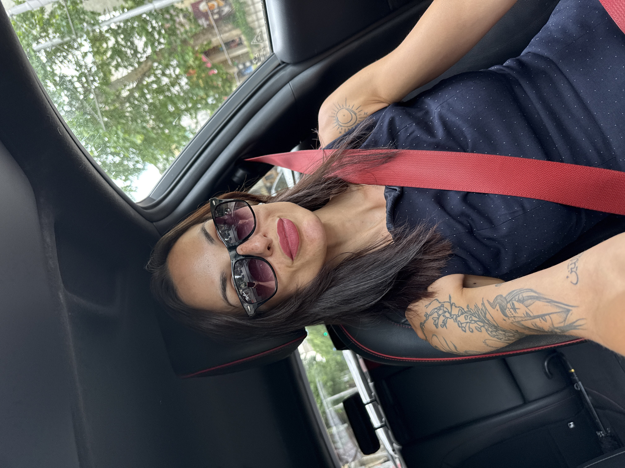 A woman is photographed from an overhead perspective while sitting in the passenger seat of a car. She is wearing a dark blue polka dot shirt and a red seatbelt. She has sunglasses on, dark hair, and red lipstick. Tattoos are visible on her arms. The interior of the car is black. Outside, blurred trees and buildings are seen through the car's window. The location is inferred to be Pleven, Bulgaria, based on the prompt.