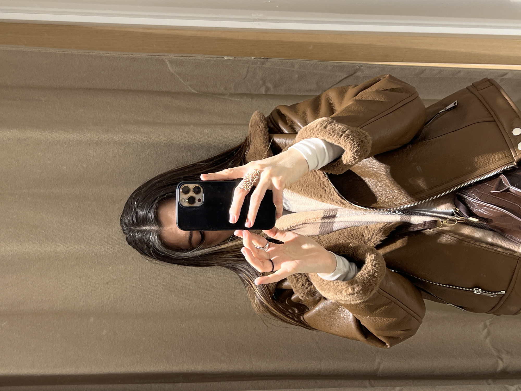 A female individual is depicted taking a mirror selfie in an indoor commercial setting, specifically a fitting room, in Riga, Latvia. She is shown from the chest up, obscuring her face with a black smartphone held in her right hand. Her left hand is raised, displaying a thin black ring on her index finger and a silver-toned, intricate filigree ring on her middle finger. She wears a brown faux leather aviator-style jacket with light brown faux shearling lining visible on the collar and cuffs, over a white long-sleeve top. A plaid scarf in shades of brown and beige is visible under the jacket. A dark brown, textured handbag with a gold-colored clip is partially visible, hanging over her left shoulder. The background consists of a light brown textured fabric curtain, typical of a changing room, with a light wood mirror frame and a white vertical surface visible on the right side.