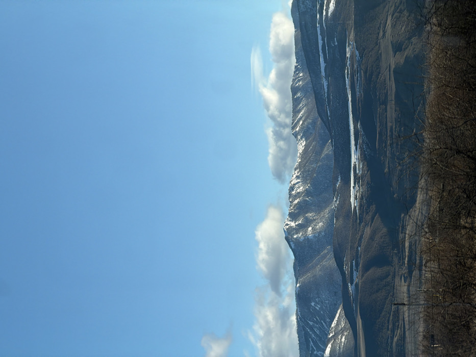 The image depicts a panoramic winter landscape in Ruisi, Georgia. The upper half of the frame is dominated by a clear, bright blue sky with scattered white cumulus clouds horizontally aligning with the mountain range. Below, multiple layers of mountains and hills stretch across the scene. The most distant mountains, occupying the mid-ground, feature prominent snowcaps on their peaks and upper slopes, contrasting with the darker, exposed rock or forested areas below. Closer hills are mostly dark brown or deep green, with some intermittent patches of snow visible in depressions or on shaded sides. The immediate foreground consists of dark, blurred, bare deciduous branches and shrubs, suggesting a lack of foliage. No people or animals are visible. No explicit actions or interactions are occurring. The environment indicates a cold, clear day. A few indistinct vertical structures, possibly utility poles or towers, are faintly visible in the lower-mid section of the landscape.