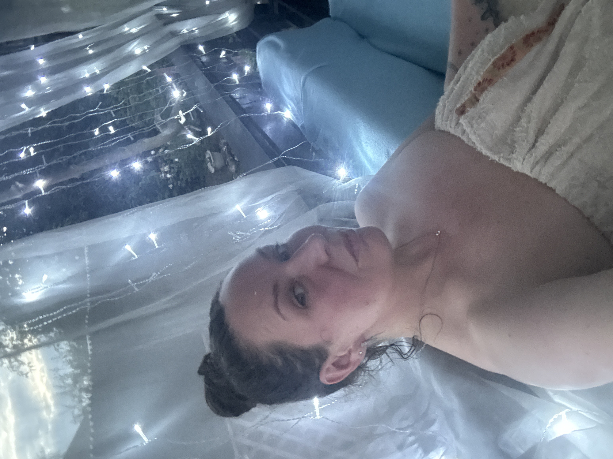 The image shows a woman wrapped in a towel, lying on a blue cushion outdoors. She has her hair in a bun and is wearing a small necklace. String lights are draped around her, creating a festive atmosphere. The background suggests an outdoor setting, possibly a backyard or patio, with trees and foliage visible through sheer curtains. The lighting is dim, with the string lights being the primary light source. The woman is looking towards the camera with a slight smile.