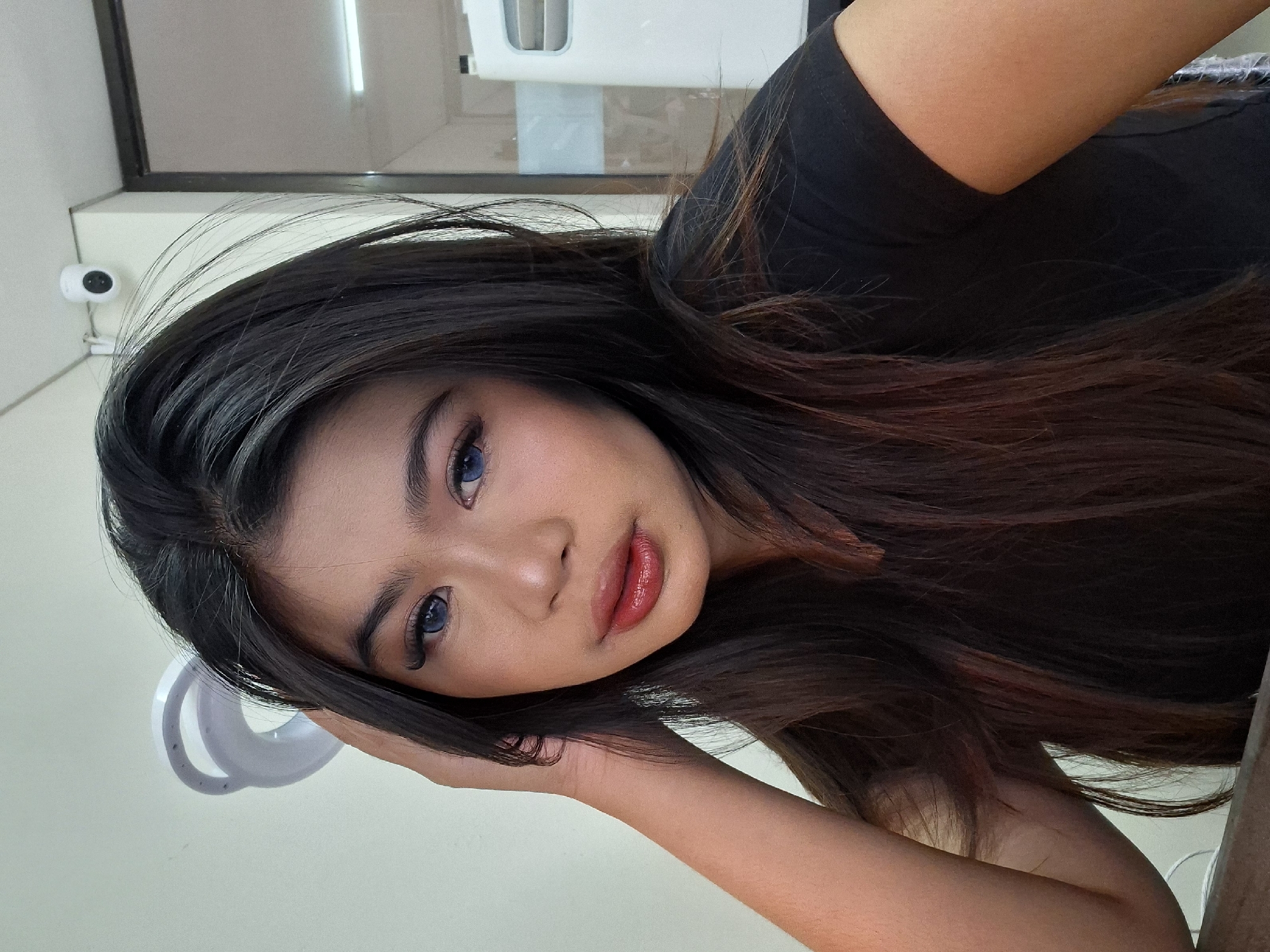 A young woman with dark hair and blue contact lenses poses for a selfie. Her left hand is raised to her head, obscuring the top left of the frame. She is wearing a black t-shirt. In the background, a white ring light is visible on the left, and a security camera is mounted on the ceiling in the upper right corner. The environment appears to be an indoor setting, possibly an office or a commercial space, with neutral-colored walls and a glass-paneled door or partition in the background. The lighting suggests an indoor setting, and there are no distinct features identifying this location as Kuta, Indonesia.