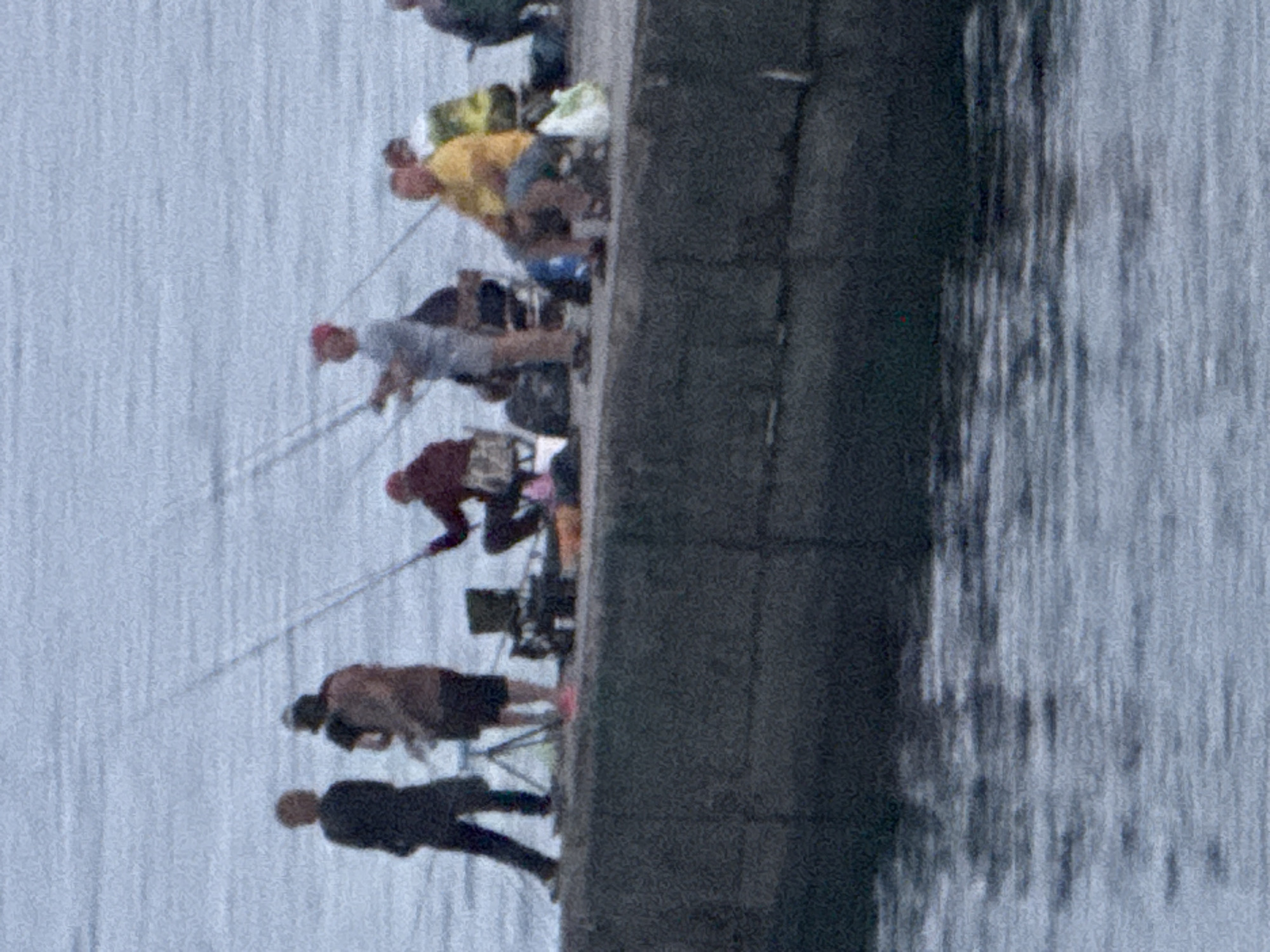 This is a photograph of a group of people fishing from a concrete pier on a cloudy day. The water appears choppy. The pier is dark grey and appears to be constructed of rough concrete. On the pier, from left to right, are: a person in a dark coat and pants, standing and holding a fishing rod; a person in a brown shirt and dark shorts, also standing and holding a fishing rod; a person in a burgundy jacket and dark pants, sitting on a red and white chair and holding a fishing rod; a person in a grey shirt and blue shorts, standing and holding a fishing rod; a person in a yellow shirt and blue shorts, sitting and holding a fishing rod; a person wearing a green jacket, partially visible, and a person in a yellow and green checkered shirt, sitting. There are bags and equipment scattered on the pier around the people. The location is identified as Partenit, Ukraine.