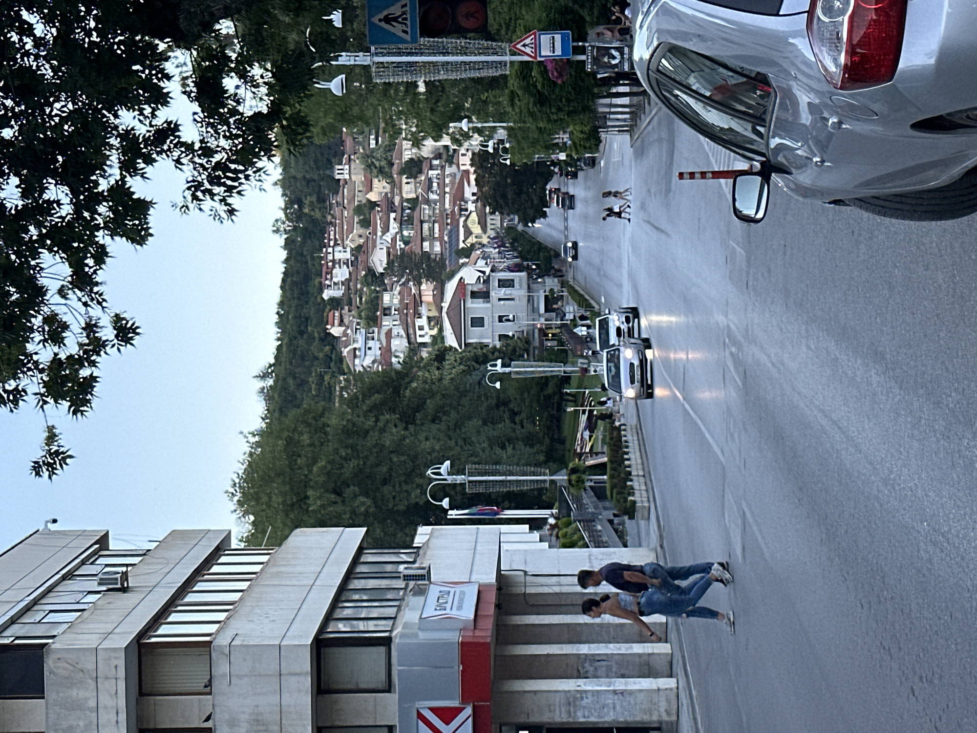 The image captures a street scene in Veliko Tarnovo, Bulgaria. In the foreground, a silver car is parked on the right side of the road. The road itself is asphalt, with visible lane markings. Two individuals, a woman and a man, are walking across a pedestrian crossing in the middle of the road. The woman is wearing ripped jeans and a light-colored top, while the man is in dark clothing. Further down the street, several other cars are driving or parked.
On the left side of the street, there is a multi-story building with a concrete facade and numerous windows. A sign with red and white lettering is visible on the building. In the background, a densely populated hillside is covered with buildings, many featuring red-tiled roofs. The architecture suggests a historic European town. Green trees are interspersed throughout the scene, with some branches from a large tree extending into the upper right corner of the frame. Streetlights and traffic signals are also present. The sky is overcast with a pale blue hue. The overall impression is that of a bustling urban environment during daylight hours, with a slight haze.