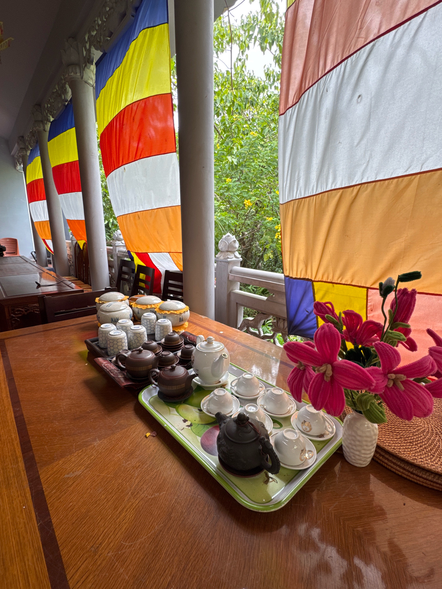 A table is set with a tea set on a tray. The tea set includes teapots, cups, and saucers. There are also several ceramic jars with lids. In the background, there are Buddhist prayer flags hanging. The flags are colorful, with blue, yellow, red, white, and orange stripes. The scene appears to be outdoors, possibly on a porch or balcony, with lush green trees and foliage visible. The wooden table has a rich, dark finish. A white vase with pink flowers sits on a woven mat to the right of the tea set. The overall setting suggests a place of worship or meditation. The presence of prayer flags strongly indicates a Buddhist temple in Vietnam, and given the tropical foliage, a location like a temple in or near the city of Nha Trang is plausible. No people are visible in the image.