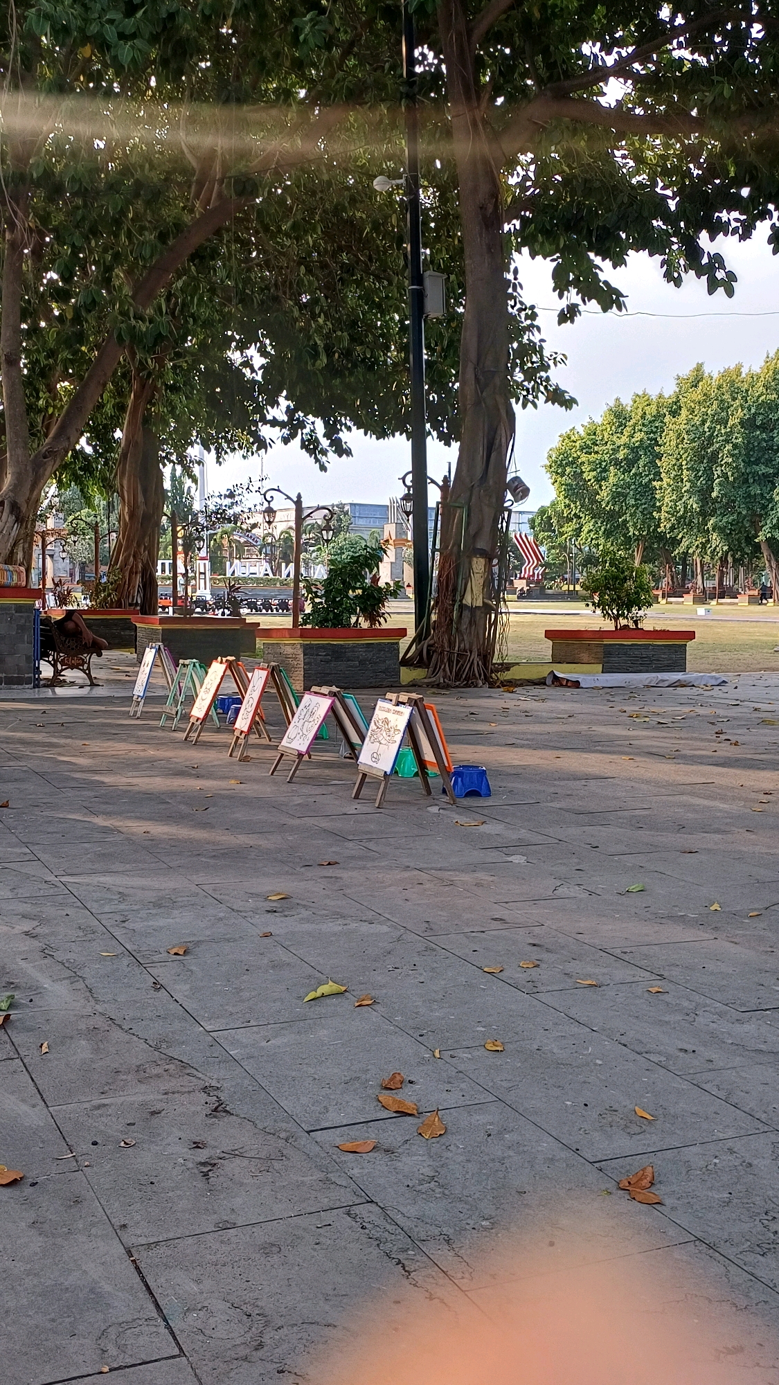 In the city of Klaten, Indonesia, a public area with a paved walkway and grassy areas features a row of small easels set up for art. Each easel displays a drawing, likely by children. The easels are of varying colors, including white, pink, green, and orange. In front of some easels are small, blue plastic stools. The scene is set under the shade of large trees with visible roots, and sunlight filters through the leaves, creating a dappled effect. In the background, there are planters with greenery, streetlights, and some buildings. Fallen leaves are scattered on the paved ground.
