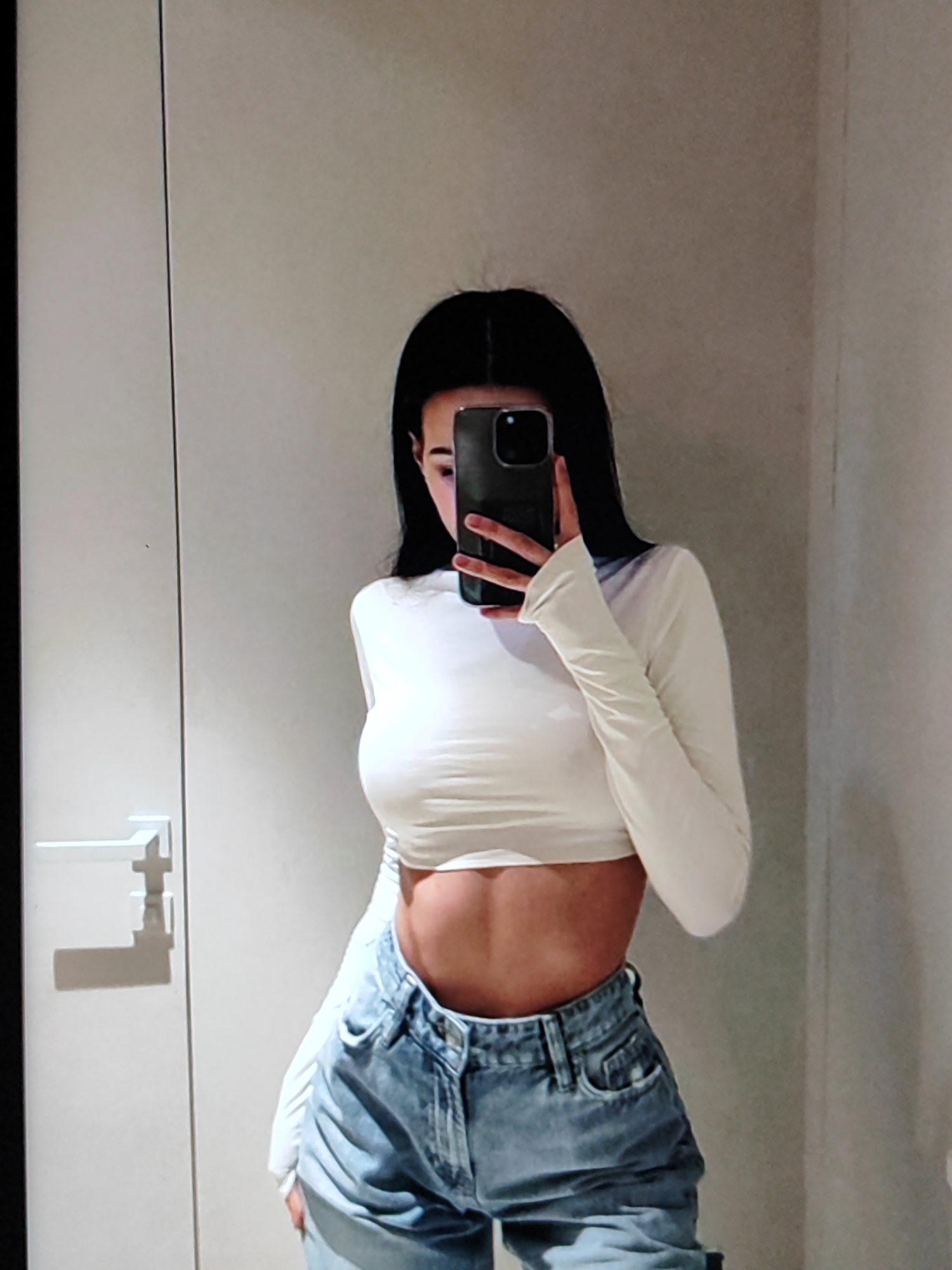 A young woman with long, dark hair takes a mirror selfie in what appears to be a room with light-colored walls. She is wearing a white, long-sleeved, cropped shirt that exposes her midriff and light-wash, high-waisted jeans. She is holding a smartphone in her right hand, obscuring her face. The room has a minimalist aesthetic with a door and a door handle visible on the left side of the frame. The lighting suggests it is daytime, and the setting is likely indoors, possibly a bedroom or dressing area. No text is visible in the image. The overall mood is casual and self-aware.