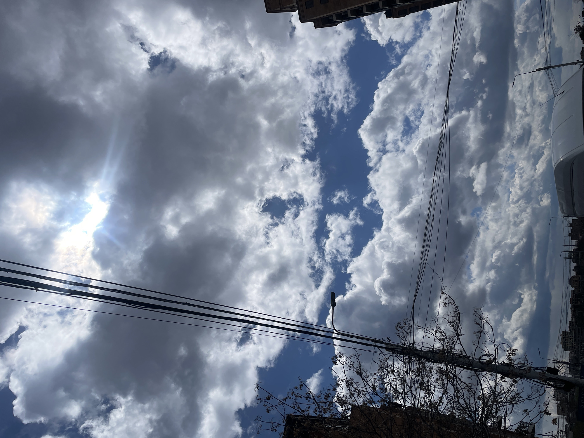 The image shows a view looking upwards towards a sky filled with cumulus clouds, with patches of blue visible in between. A bright light source, likely the sun, is obscured by clouds in the upper left. Several power lines stretch across the lower half of the frame, obscuring portions of the sky and clouds.
In the lower right corner, there are leafless branches from a tree, with the corner of a dark-colored building behind them. Faintly visible on the right edge of the frame is a vehicle. Barely visible buildings at the very top and very bottom right, indicating an urban environment.
Based on the architecture and overall scene, the location is Yerevan, Armenia. There are no people visible in the scene. The focus is on the sky, clouds, and utility infrastructure within a city setting.