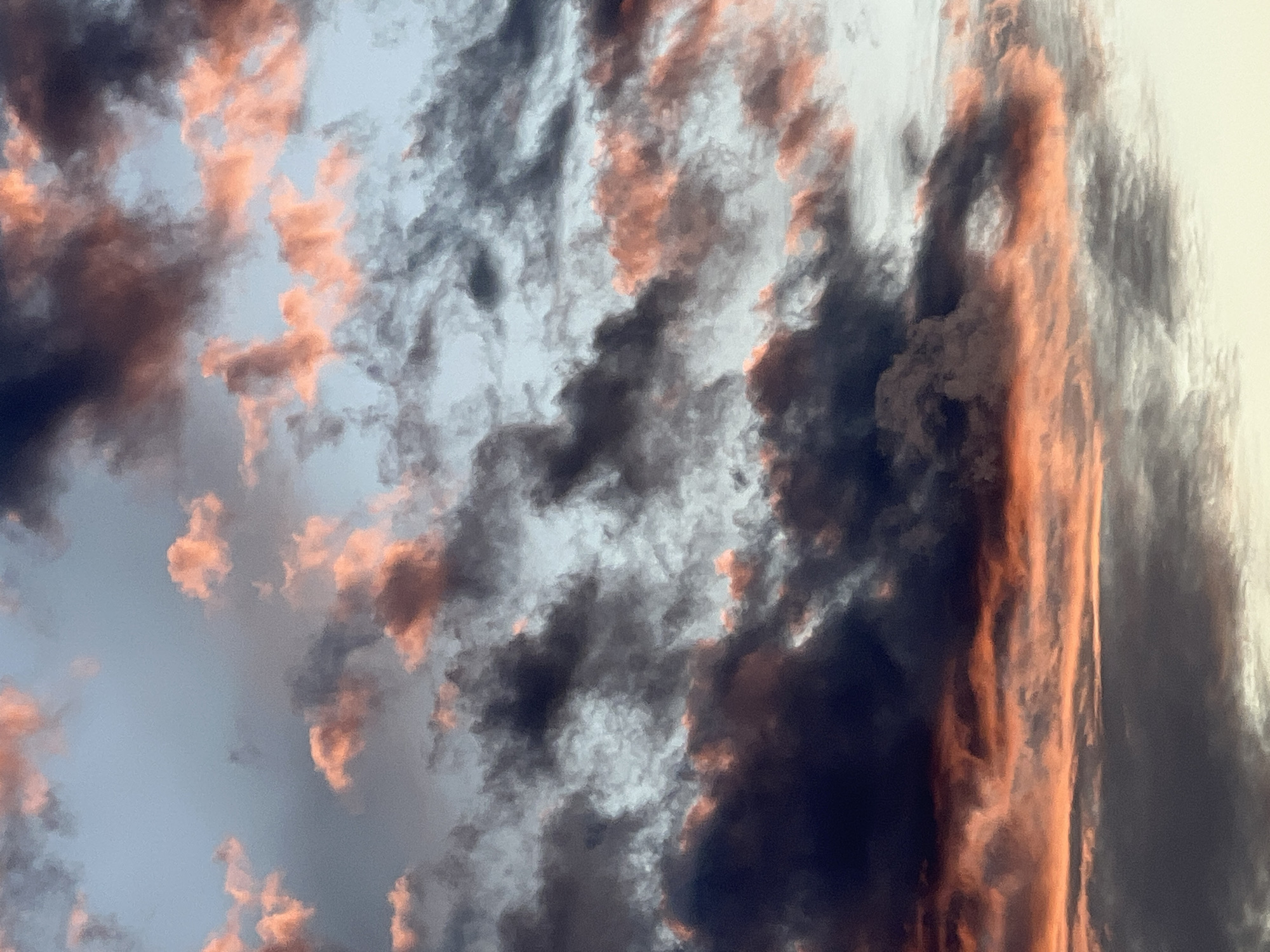 The image depicts a dramatic sky filled with dark, swirling clouds tinged with vibrant hues of orange and pink, suggesting a sunset or sunrise. The colors are intense, with deep shadows contrasting against the illuminated portions of the clouds. The clouds appear thick and textured, with some areas catching the light more brightly than others, creating a sense of depth and movement. The overall impression is one of awe and perhaps a touch of foreboding due to the dark tones, but the warm colors also bring a sense of beauty. There are no discernible people, objects, or text in the image. The scene is entirely focused on the celestial display. The weather appears to be overcast with dramatic cloud formations illuminated by low-lying sun. The location context provided is Sinyavino, Russia.