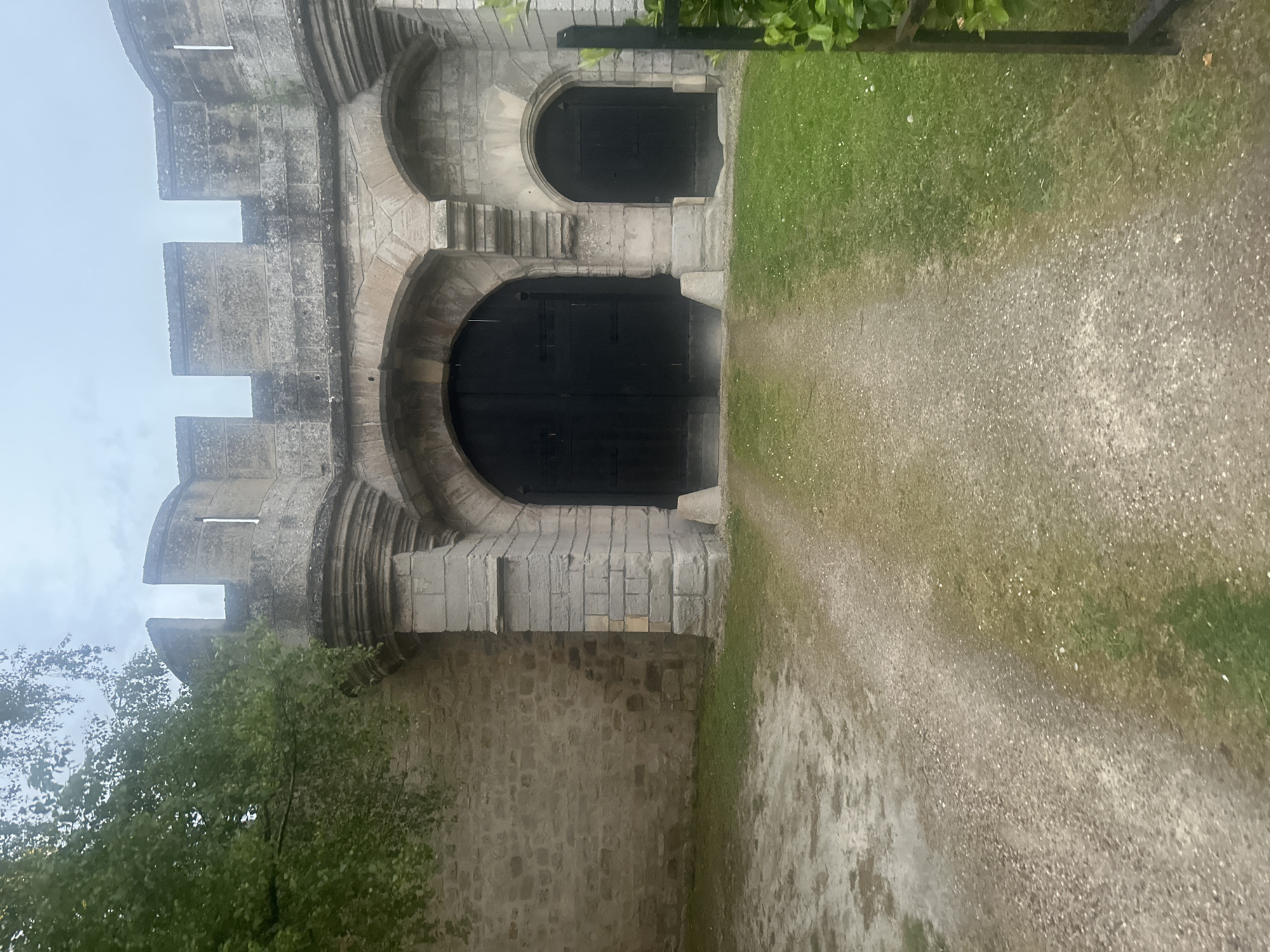 The image captures a historic stone building, likely a castle or fortified abbey, viewed from a ground-level perspective. The structure, made of light-colored, weathered stone blocks, dominates the left side of the frame, showcasing robust architectural details. Its upper section features crenellated battlements and narrow, rectangular openings or loopholes.
Two prominent arched doorways, each fitted with heavy, dark wooden doors, are visible. The larger arch is positioned lower and more centrally within the building's facade. Above and slightly to its right, a smaller arched doorway with a similar dark wooden door is set at a higher level, accessed by a series of stone steps built into the wall. The stone facade displays natural texture and signs of aging.
To the right of the building, a light-colored path, appearing to be made of dirt or gravel, extends into the distance. Adjacent to the path, patches of green grass with scattered lighter specks (possibly small flowers or fine gravel) cover the ground. Green foliage from trees or bushes is visible in the lower left foreground and extends into the upper right corner of the image.
The sky is uniformly overcast and light gray, indicating a cloudy day with soft, diffused lighting. There are no harsh shadows, suggesting an absence of direct sunlight. No people or specific activities are depicted, and no visible text is present. The scene evokes a sense of history and permanence. This location is in Saint-Leu-d'Esserent, France.