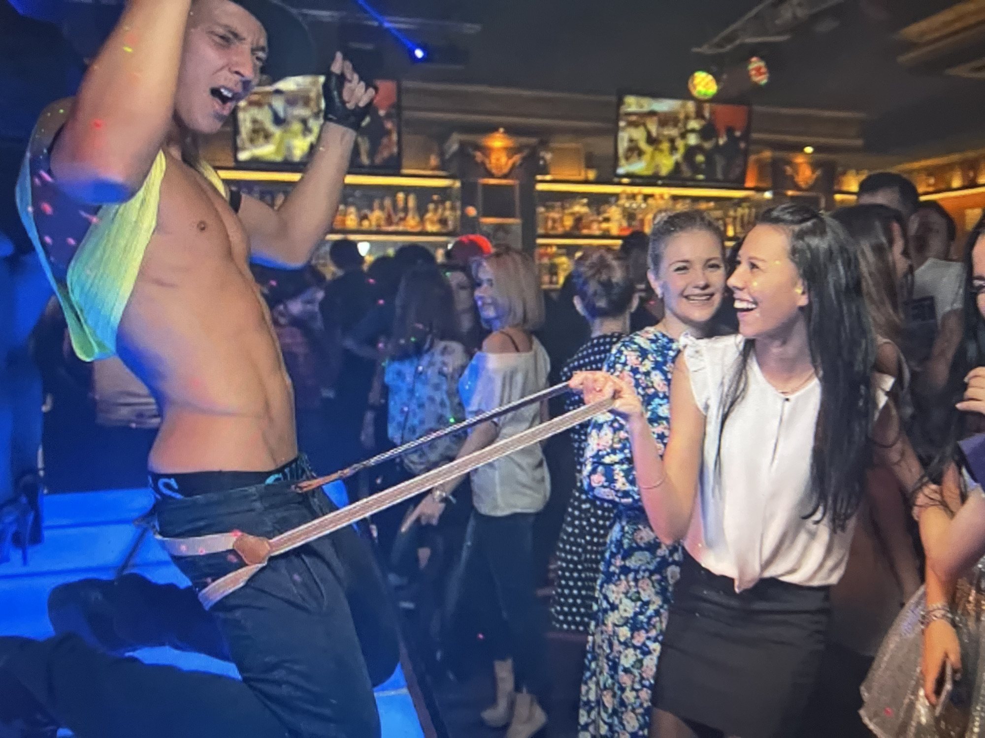An indoor scene, possibly a nightclub or bar, is depicted. The foreground features a male performer, shirtless and wearing a black hat and pants, engaged in an action that involves a light-colored garment. He appears to be performing or dancing, with his mouth open in an expression. Behind him, a group of women are visible, some smiling and looking towards the performer, while others are partially obscured. The background shows a bar area with shelves stocked with bottles and a television screen. The overall lighting is dim, with some colorful lights casting a glow on the scene. The setting appears to be in the city of Qarağandı city administration, Kazakhstan.