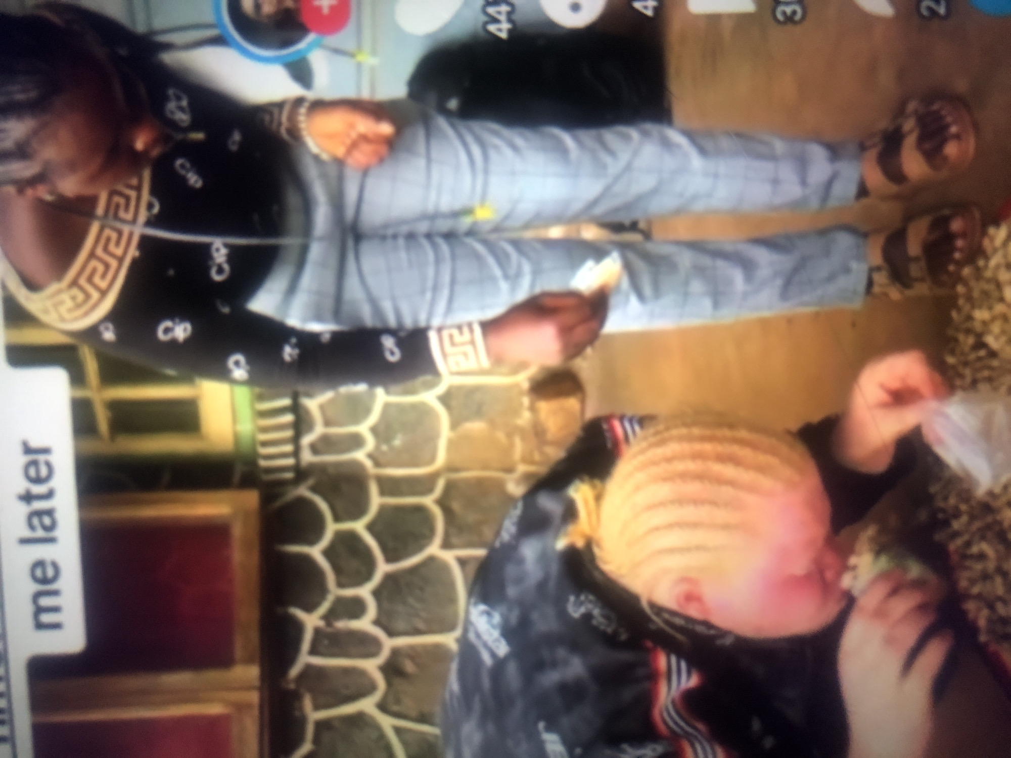 The image shows two people, one of whom is a child with albinism. The child, lying on the ground, is leaning over to examine a pile of small, dark objects and a plastic bag. The other person, an adult wearing a black long-sleeved shirt and checkered pants, is standing and looking down at something in their hand. The adult is holding what appears to be money. The background suggests an outdoor setting with stone wall detailing and wooden window frames, possibly in a residential area of Howayi, Nigeria. A text overlay on the top of the image reads "me later." The overall scene depicts a moment of observation and potential exchange or interaction.