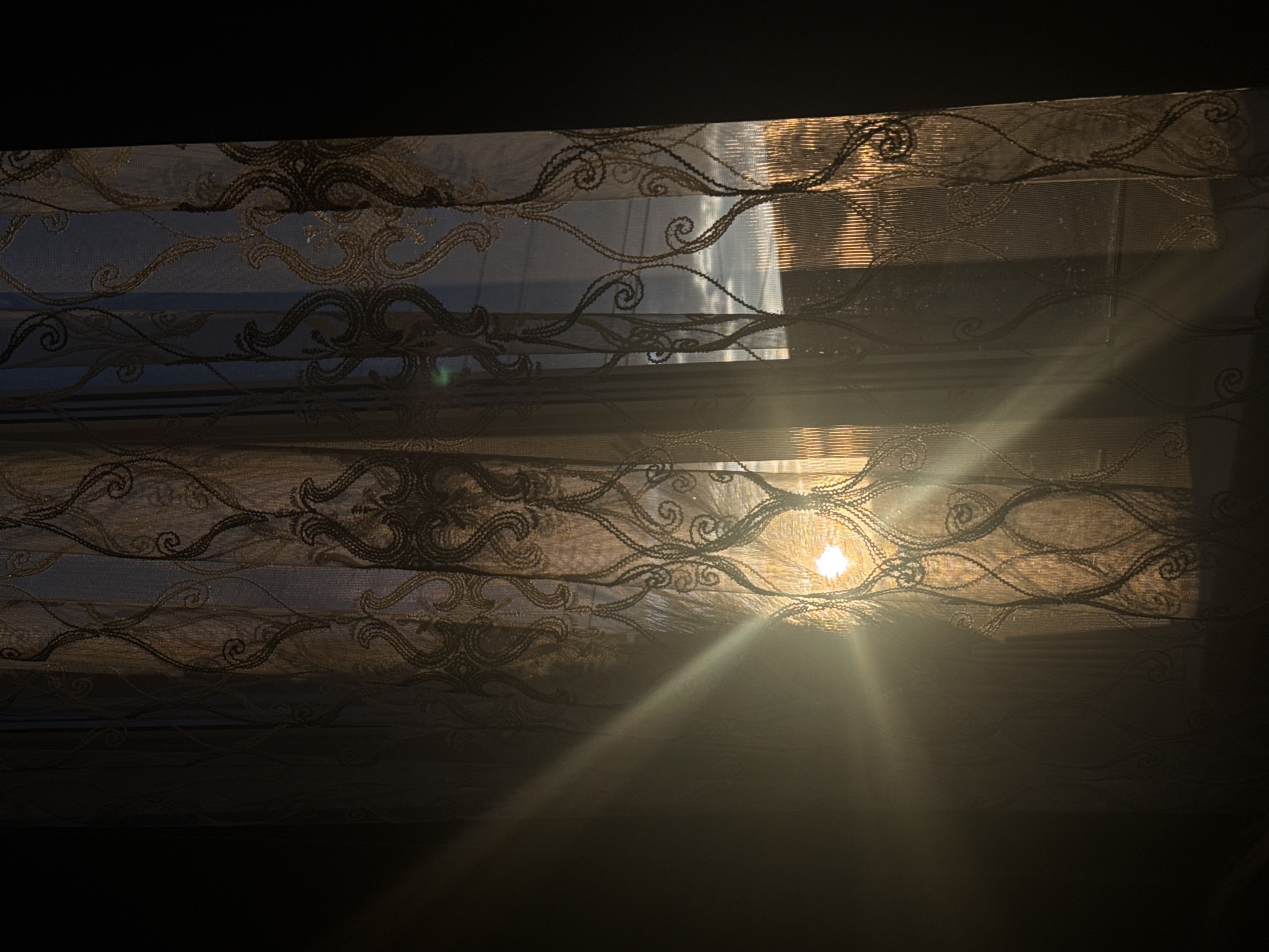The image depicts a sunbeam entering a room through a patterned sheer curtain. The curtain is translucent, allowing the sunlight to filter through and cast a warm glow on the left side of the frame, creating a hazy effect. The sun itself is visible as a bright, almost white circle behind the curtain, emanating strong rays of light. The curtain has an ornate, swirling pattern in dark thread. Through the curtain, a glimpse of the outdoors can be seen, showing a sky with clouds and what appears to be a cityscape or buildings in the distance, illuminated by the setting sun. The right side of the image is darker, with the curtain and the window frame creating a dark silhouette against the brighter, sunlit portion. The scene appears to be an interior shot taken in Rayevskiy, Russia, during either sunrise or sunset.