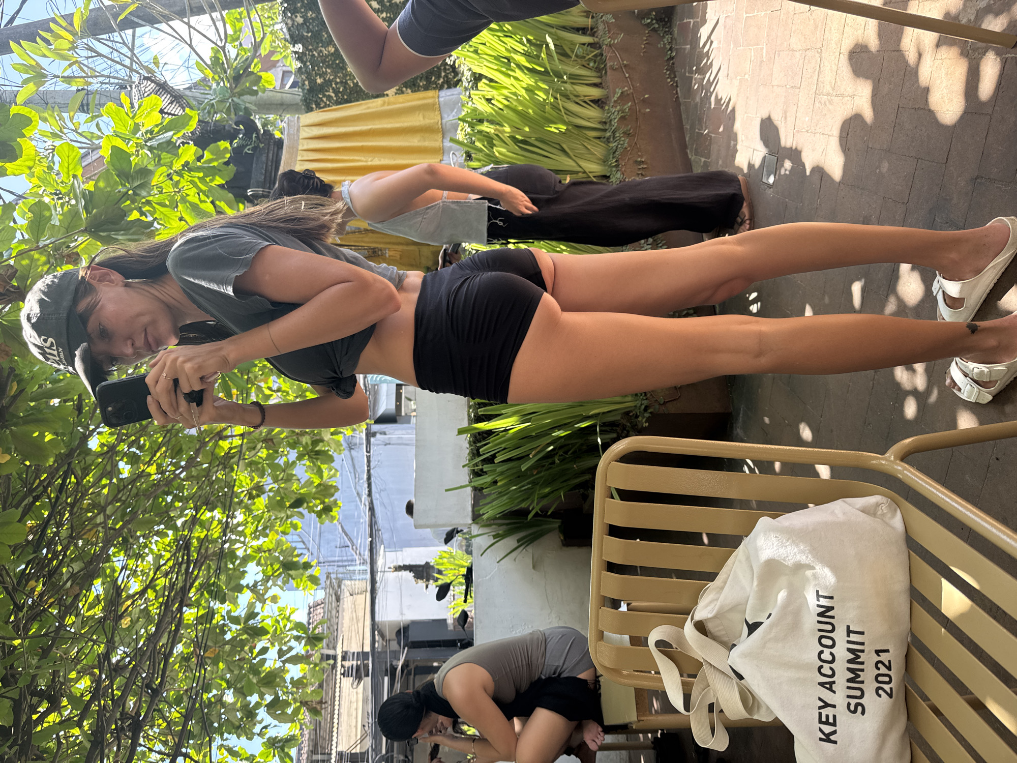 The image, taken in the city of North Kuta, Indonesia, shows a person holding a phone as if taking a photo or video. The person is wearing a t-shirt, athletic shorts, and slides, and a hat, and appears to be standing outside in an urban area with buildings, trees, and other people visible. Other people are on the scene. The bag on the chair is marked "Key Account Summit 2021".