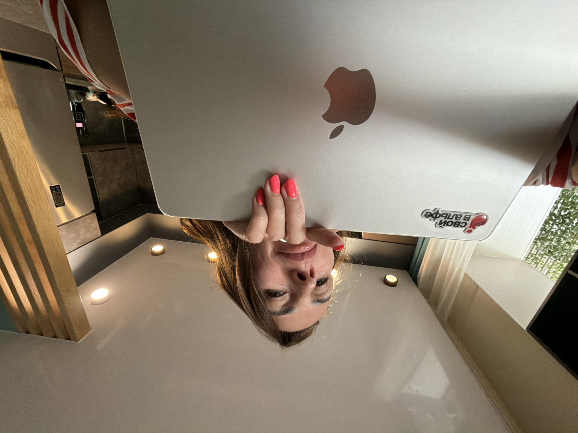 This image depicts a woman holding an open laptop, positioned in a modern interior setting in Birobidzhan, Russia. The perspective is low-angle, looking upwards at the woman who peers over the top edge of the laptop screen directly at the viewer.
The central subject is a woman with long, dark brown hair and a light complexion, appearing to be in her 30s or 40s. Her face, partially illuminated by ambient light, shows subtle eye makeup and full, slightly parted lips. Her left hand is visible on the top edge of the laptop, displaying bright pink-orange nail polish on all visible fingers. She wears a garment with red and white stripes, visible on her right side beneath the laptop.
The laptop is a silver-colored Apple MacBook, held horizontally. The iconic Apple logo is prominently displayed on the lower right portion of its lid. On the upper left of the lid, a red and white sticker is affixed, featuring Cyrillic text that reads "Свой в Альфе" (Svoy v Alfe), translating to "One's own in Alfa," suggesting an affiliation with Alfa-Bank.
The background reveals a contemporary interior space, likely a kitchen or open-plan living area. To the left, a window provides natural light, showing green foliage outside, partially obscured by a sheer white curtain. The ceiling is white and features multiple recessed circular light fixtures, casting a warm glow. To the right of the woman, a tall, silver refrigerator is visible. Adjacent to the refrigerator, a decorative vertical wooden slatted panel extends from the floor or counter upwards. Behind these elements, a dark, possibly textured wall or cabinetry is present, with another light fixture visible in the upper right. A small, dark electronic device is discernible on a counter surface behind the laptop, between the dark wall and the refrigerator. The overall lighting is a combination of natural light from the window and artificial illumination from the ceiling fixtures.
