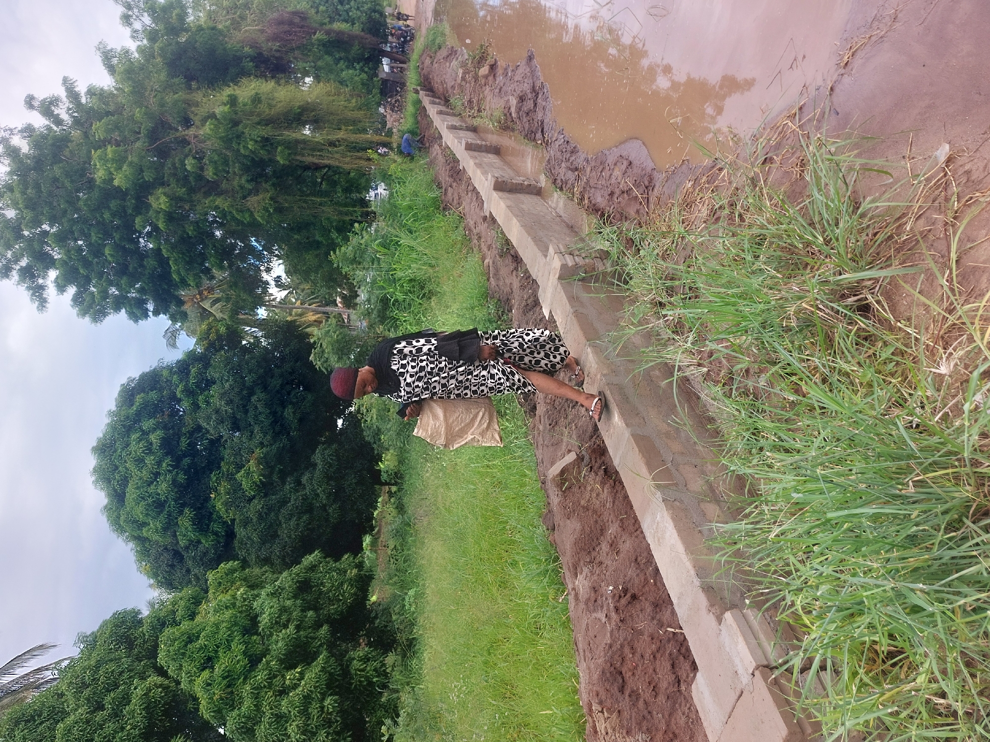 A woman walks along a low, concrete canal beside a grassy, muddy area. The canal appears to contain stagnant, brown water in sections. The woman is wearing a dark patterned dress, a head covering, and sandals, and she carries a light-colored bag. Lush green trees and vegetation dominate the background. The picture's location is likely in Dar es-Salaam, Tanzania, based on the environment and style of the canal and clothing.
