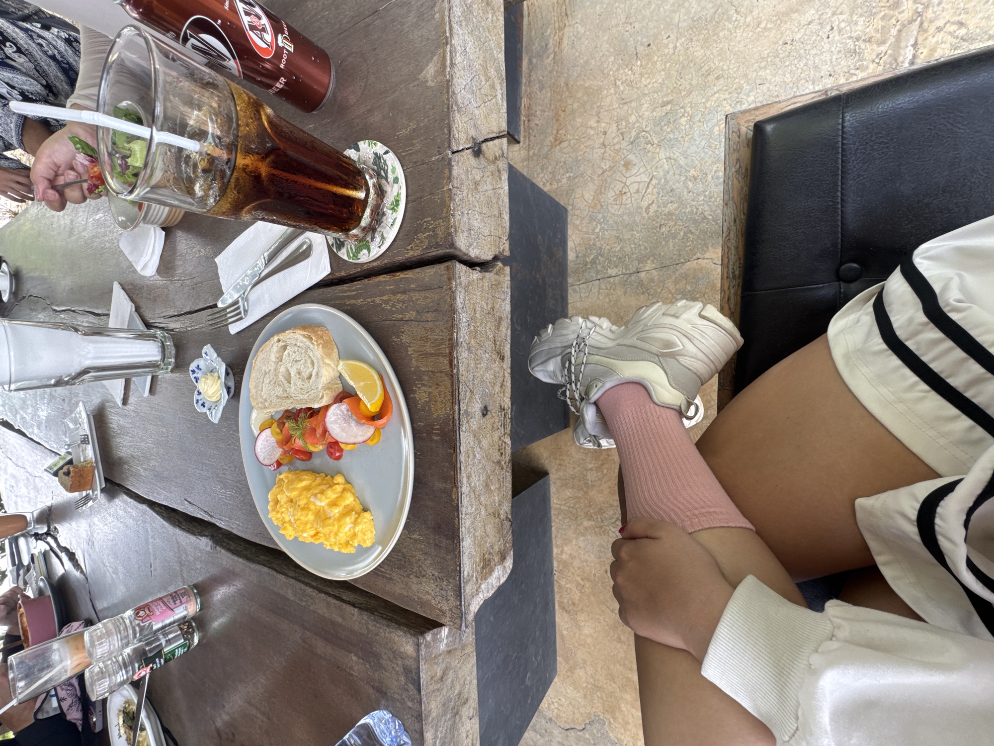 The image is a high-angle, close-up shot of a meal scene with a person's leg visible. The location is purportedly Ban Tom, Thailand. The scene features a wooden table with a plate of food, including bread, eggs, and sliced vegetables. There is also a tall glass of dark liquid with ice, a bottle of root beer, and seasoning shakers. A person's arm and hand are visible near the glass, holding a fork, and another person's arm is visible. Beside the table, a person sits on a black cushion chair. The person is wearing a pink sock and white sneakers. They also have on shorts and a white long-sleeved shirt. One hand is visible near the knee. The overall composition suggests a casual dining setting.