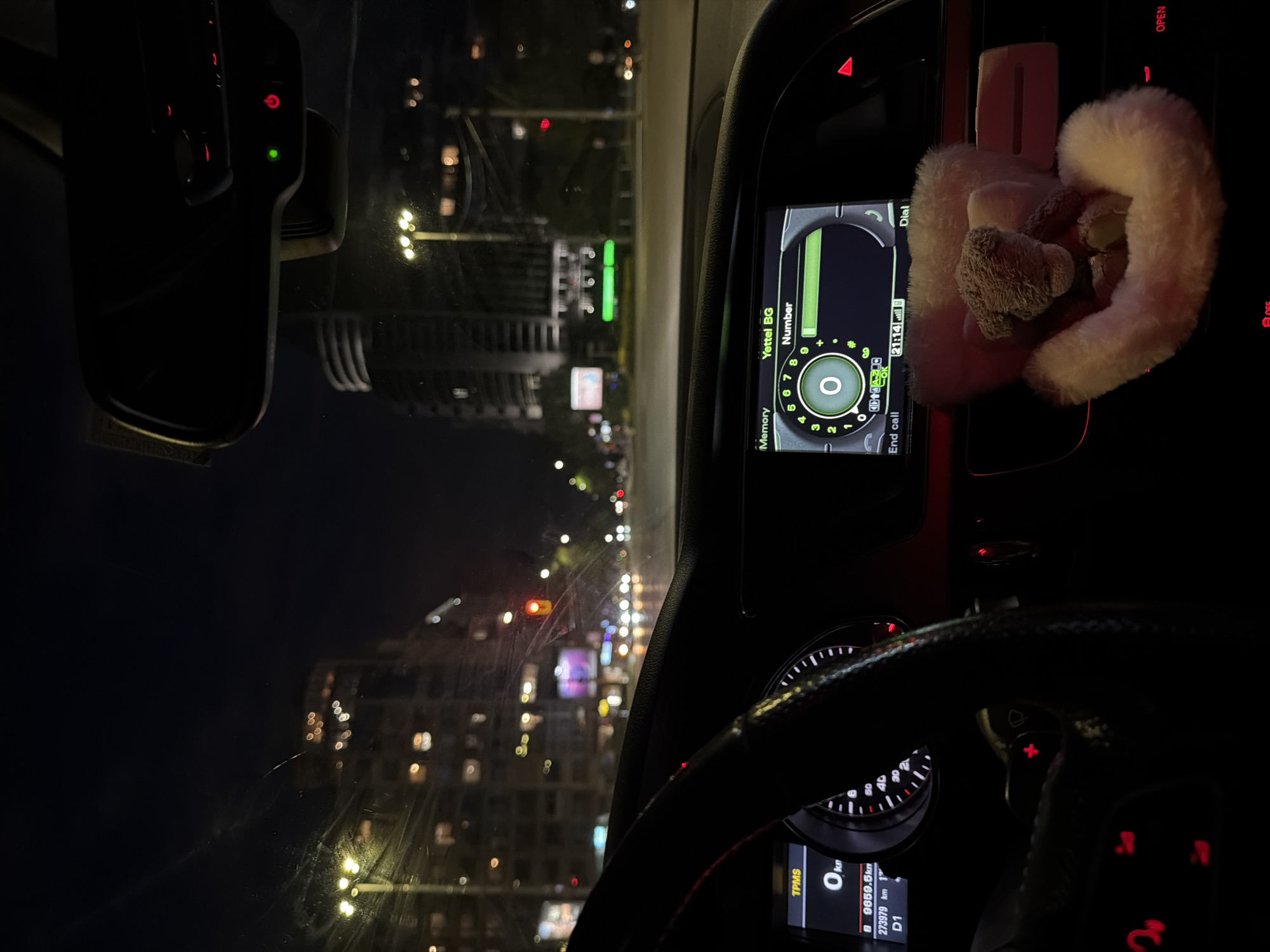 This vertical, nighttime image is captured from inside a car, looking out through the windshield at a city scene. The interior of the car occupies the right side of the frame, showcasing parts of the dashboard, infotainment system, and steering wheel.
On the car's infotainment screen, a phone dialer interface is visible. It prominently displays "Yettel BG" and "Number," with a large "0" in the center of the dial pad. Other text on this screen includes "Memory," "Dial," and "End call." The time "21:14" is also present on the screen. Hanging from the dashboard, adjacent to the screen, is a fluffy, light-colored car accessory, which appears to have a small, plush teddy bear attached. Further down, parts of the steering wheel and instrument cluster are visible, displaying information such as "TPMS," "0 km," "9659.5 km," "273979 km," and "D1," along with speedometer markings. Various buttons with red backlighting are seen on the dashboard and steering wheel. In the upper left corner, the rearview mirror is partially visible, showing small red and green indicator lights.
Through the windshield on the left side of the frame, a blurry cityscape unfolds at night. Numerous illuminated windows in distant buildings and bright streetlights create a soft bokeh effect. Red and white lights from other vehicles or traffic signals are also discernible. The scene suggests the car is either stationary or moving slowly in an urban environment during the late evening. The visible text "Yettel BG" provides a clear location cue, indicating the setting is in Bulgaria, consistent with Sofia.