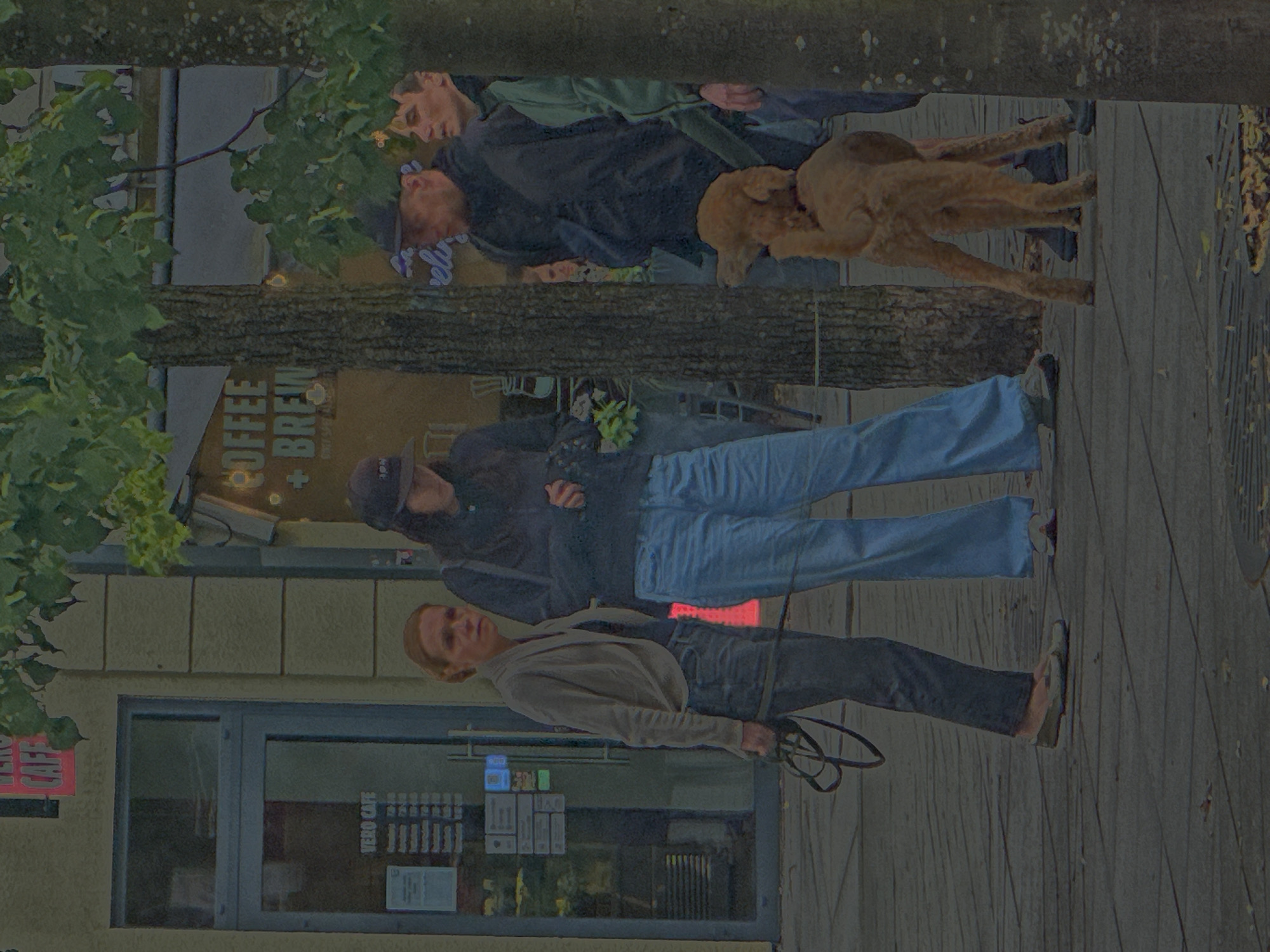The image depicts a group of people and a dog on a sidewalk in Kaunas, Lithuania, in front of a building with a cafe. A woman in a light-colored jacket and dark pants stands on the left, holding a dog leash. Beside her, a person wearing a black baseball cap and blue jeans is partially obscured. Further to the right, two men and another person, possibly a child, are visible. One man wears a black jacket and a baseball cap, while the other, in a green jacket, is seen from the side. A light brown poodle is standing in the foreground, looking to the left. Behind the group, a tree trunk is prominent. The building features a glass door with "VERO CAFE" signage and a list of operating hours displayed. Another sign on the building reads "COFFEE + BREW." The ground is paved with grey cobblestones, and fallen leaves are scattered on the surface. The overall lighting suggests an overcast day.