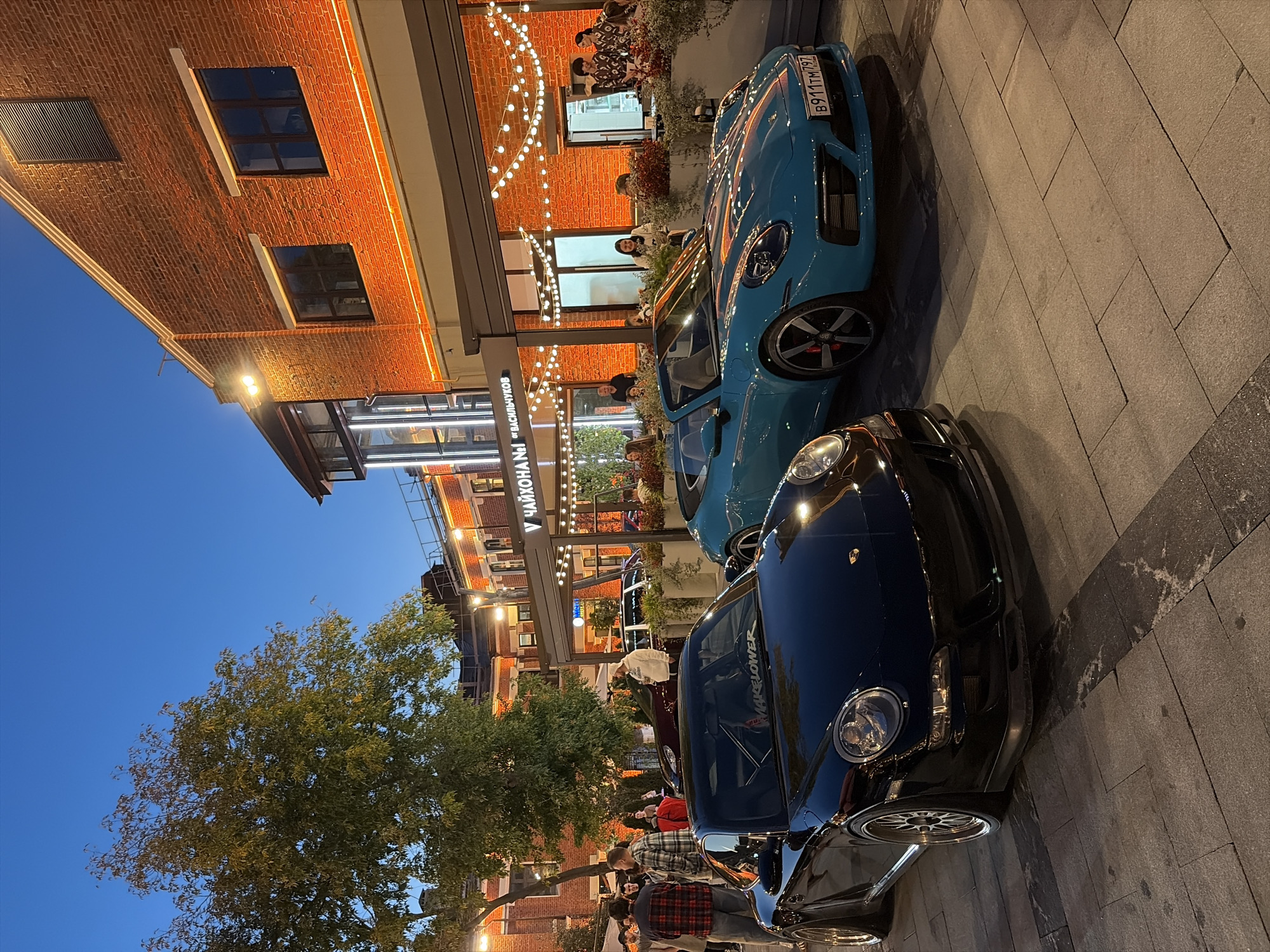 At dusk in Moscow, Russia, two high-performance sports cars, a black Porsche and a teal convertible Porsche, are parked on a cobblestone walkway in front of a brick building adorned with string lights. A group of casually dressed individuals stand near the black Porsche, some conversing while others observe the vehicles. The teal Porsche, a convertible, is positioned to the right of the black one. The building appears to be a restaurant or cafe, with patrons visible inside and seated at outdoor tables. A large tree with green foliage is situated to the left of the frame, contributing to the urban outdoor ambiance. The lighting suggests twilight or early evening, with artificial lights illuminating the building and surrounding area.