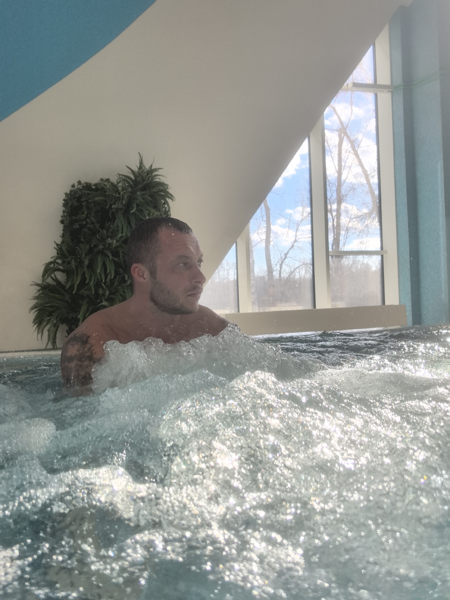 A fair-skinned man is chest-deep in water, likely a hot tub or whirlpool, with strong bubbles. He has short, dark hair, a beard, and a tattoo on his left upper arm. He looks to his right, away from the camera. Behind him is an artificial plant. Natural light streams in from a triangular window showing bare tree branches against a blue sky with clouds. The architecture has light blue and white accents. Based on visual evidence, this might be located at the Quaris Spa & Hotel in Nizhny Novgorod, Russia, as its interior features a similar design.