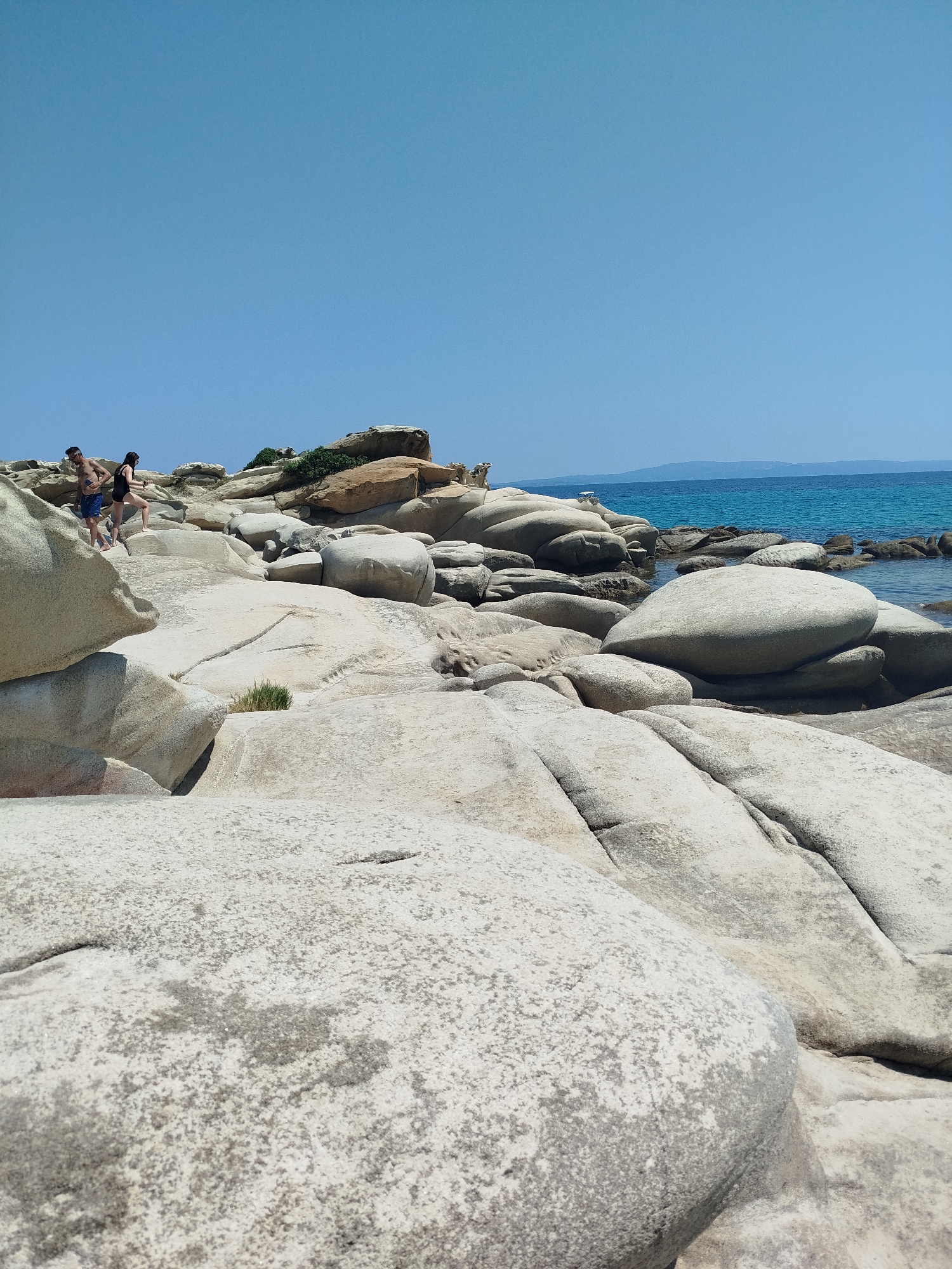 This image captures a scenic coastal view in Sithonia, Greece. The foreground is dominated by large, smooth, light-colored rocks, some of which are partially covered in a textured, granular surface. In the middle ground, two individuals, a man and a woman, are walking amongst similarly shaped rocks. The man is wearing blue shorts and the woman is wearing a dark swimsuit. They are positioned on a rocky incline. Further back, the sea is a vibrant turquoise, meeting a clear blue sky. The horizon shows a distant landmass. Several more large rock formations are scattered along the coastline, with some vegetation visible on the higher rock ledges. The overall impression is one of a natural, sun-drenched beach environment.
