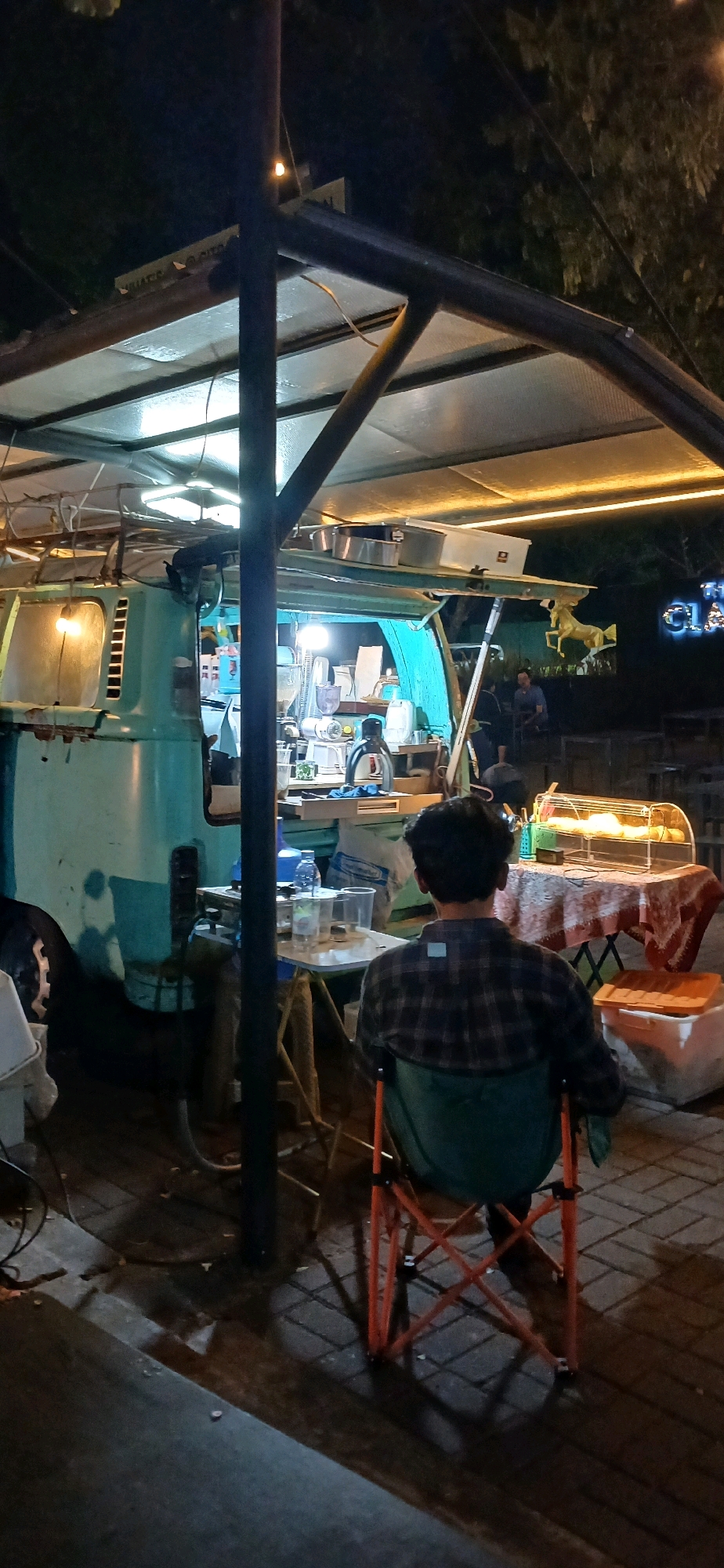 The image depicts an outdoor scene at night, likely a food stall or cafe. In the foreground, a person wearing a plaid shirt is seated on a folding chair, facing away from the camera. Behind them, a teal-colored van serves as a food truck, with its interior illuminated and stocked with various equipment, possibly for preparing drinks or food. A table with cups and a bottle is visible near the van. Further back, there is a display case filled with food items and another table draped with a patterned cloth. In the background, a sign with illuminated letters, which appears to read "THE CLASSIC," is mounted on a building or structure. The overall environment suggests a commercial or recreational area, possibly in the city of Cirebon, Indonesia, given the context provided. The ground is paved with bricks, and trees are visible in the darkness. Artificial lighting from the food truck and string lights creates a warm, inviting ambiance.