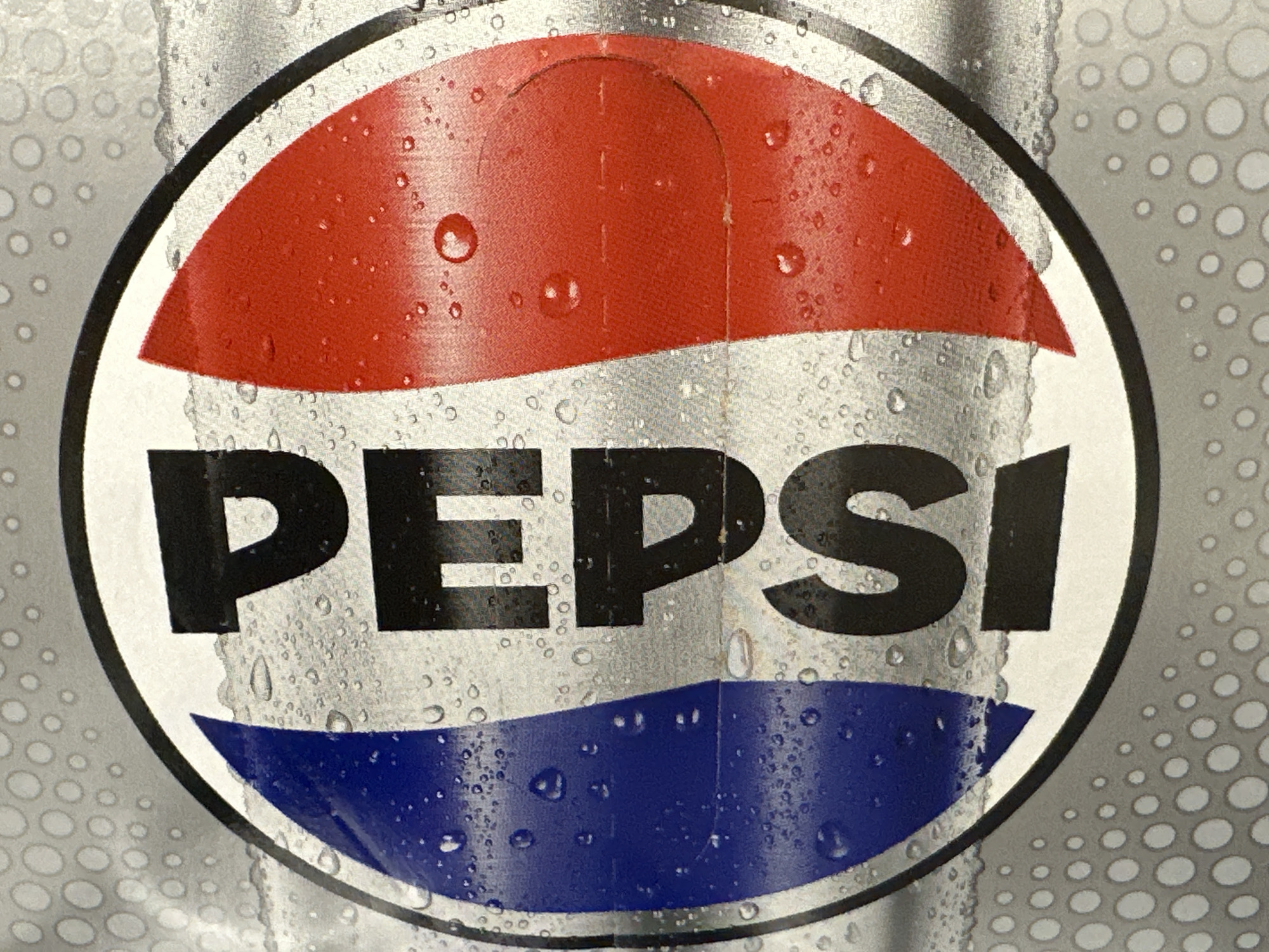 The image is a close-up, eye-level shot of a Pepsi can. The can is metallic silver with a condensation of water droplets covering its surface. The iconic Pepsi logo is prominently displayed in the center, featuring a red and white curved band on top and a blue curved band on the bottom, with the word "PEPSI" in large black letters in between. The background of the can has a pattern of small, light gray circles. The image is devoid of any contextual information regarding its specific location, time, or any human presence.