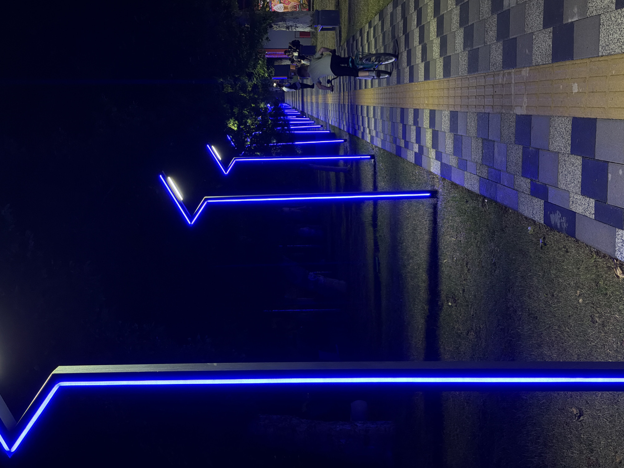 The image depicts a pedestrian pathway illuminated by a series of modern, vertical blue neon lights. The pathway itself is paved with a geometric pattern of dark gray and light gray square tiles, with a textured yellow tactile paving strip running down its center. On the right side of the pathway, a young person is riding a bicycle away from the camera. Trees line the pathway on both sides, casting shadows in the dim light. In the background, other blue lights extend into the distance, creating a rhythmic and modern aesthetic. The scene appears to be a public park or promenade in the city of Chirchik, Uzbekistan, likely at night, given the prominent artificial lighting and the dark sky.