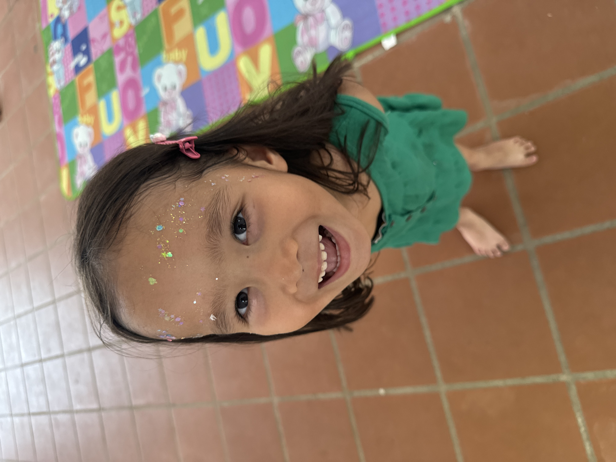 A child with dark hair and light brown skin is seen from a high-angle perspective, smiling broadly. The child has glitter on their forehead and wears a pink clip in their hair. They are dressed in a green garment. Part of a colorful alphabet and teddy bear patterned mat is visible in the background. The floor is covered in large, square, terra-cotta tiles. The location is believed to be Mũi Né, Vietnam, as this information was provided.
