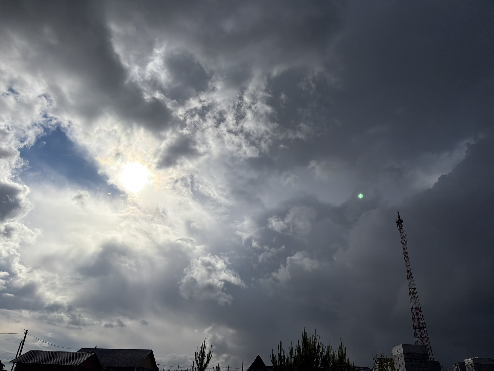 The image captures a dramatic sky dominating the frame, characterized by a complex interplay of clouds and light. On the left side, bright, white and grey clouds are partially illuminated by a visible, intense sunburst, with a sliver of clear blue sky peeking through above the sun. This contrasts sharply with the right side of the image, which is consumed by much darker, heavier storm clouds, creating an ominous atmosphere.
Extending high into these dark clouds on the right is a tall, lattice-style communication or radio tower, its intricate structure silhouetted against the somber sky. Near the top of the tower, and slightly to its left within the dark clouds, a distinct green circular lens flare or light anomaly is visible.
At the bottom left and center, the dark silhouettes of residential rooftops and power lines suggest a populated area. Sparse, dark outlines of tall, slender plants or young trees are also visible along the very bottom edge of the frame. The overall impression is of a dynamic weather event, possibly a break in a storm or an approaching one, with the sun fighting through heavy cloud cover. The location is Chaikovsky, Russia. There is no visible text in the image.