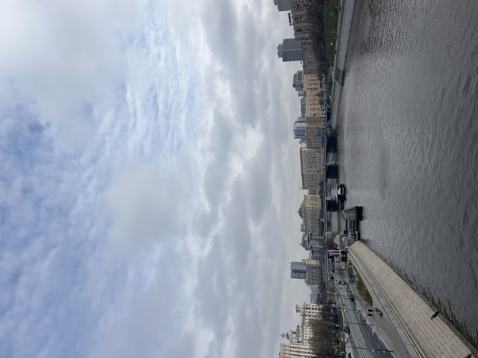 The image depicts a cityscape in Moscow, Russia, along the Moskva River. The river is on the right side of the image, showing its concrete embankment and the water's surface. A multi-lane road runs parallel to the river, with sparse vehicular and pedestrian traffic visible. The buildings lining the river include both older and modern structures, ranging from traditional Stalinist architecture to contemporary skyscrapers. The sky is overcast with a thick layer of clouds.