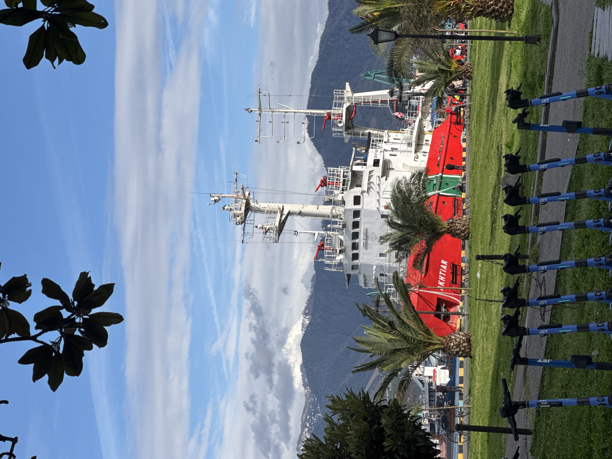 The image shows a red and white ship docked on a sunny day with a blue sky and some scattered clouds. The city and country are unknown. In the background, there is a mountain partially covered with clouds. Palm trees and other green vegetation are visible in the foreground and around the ship. A row of blue "JET" branded scooters are parked on the right side of the image. Parts of what appear to be overhead lamps are visible along a grassy area next to the scooters. Some dark foliage is visible in the top left corner of the image.