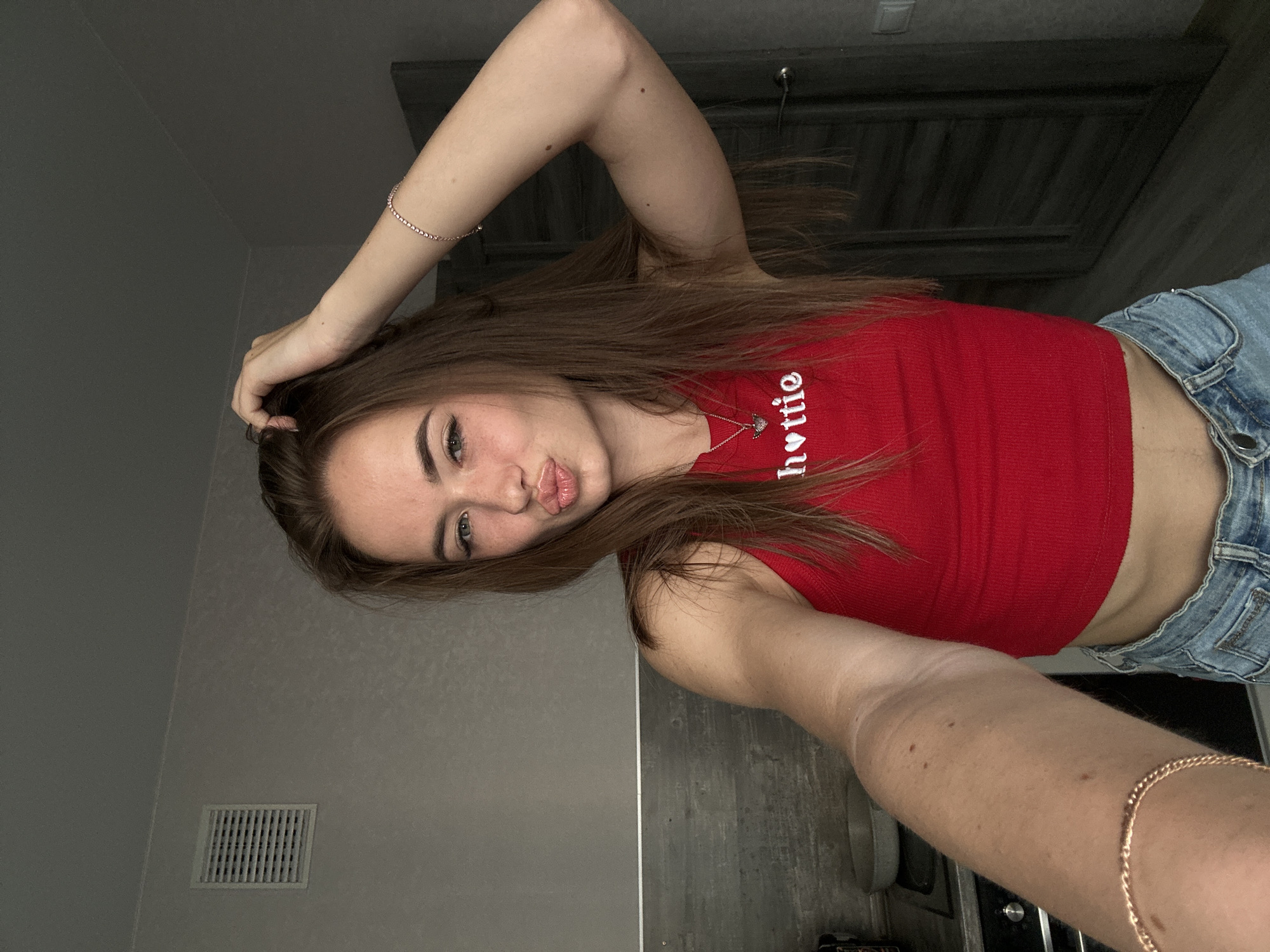A young woman with long brown hair is taking a selfie in a room. She is wearing a red tank top with the word "hottie" written on it in white letters, and denim shorts. She has her right hand on her head and is puckering her lips. A bracelet is visible on her right wrist. The background appears to be a domestic interior with a vent on the wall, a kitchen counter with a pan, and a dark door. The lighting suggests it is daytime.