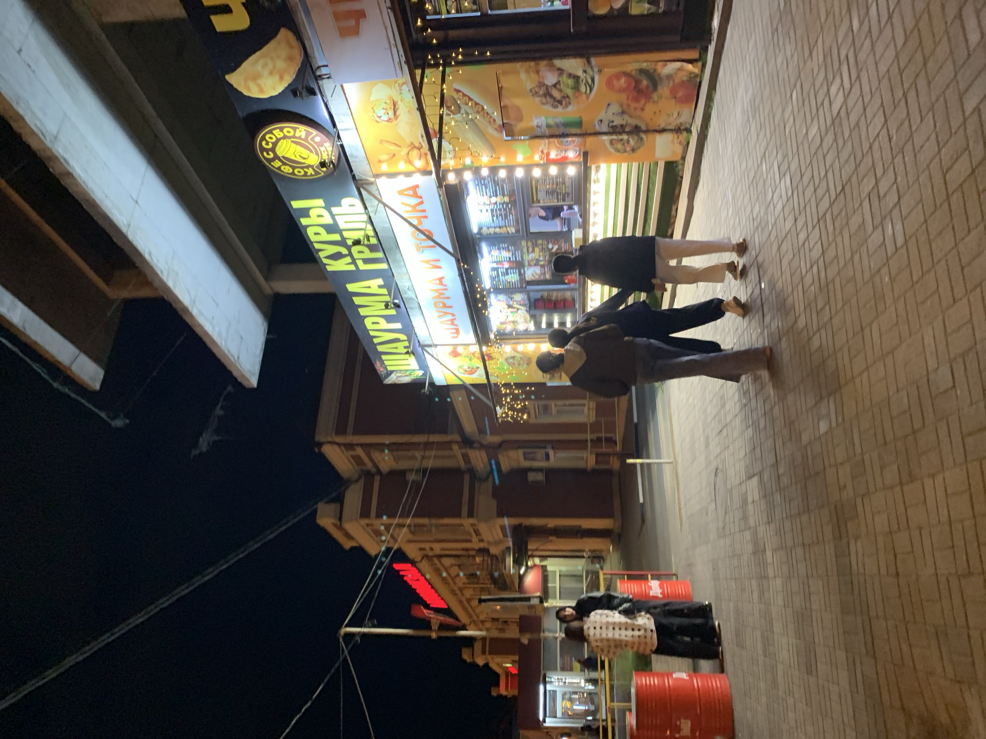 The image depicts a street scene at night, featuring a group of people walking near a food stall. The food stall displays a sign in Cyrillic script that reads "ШАУРМА И ТОЧКА" and "СОБОЙ КУРЫ ГРИЛЬ", suggesting it sells shawarma and grilled chicken. The stall is brightly lit with multiple light fixtures, and images of food are displayed. Pedestrians are walking past the storefront on a brick-paved sidewalk. Utility poles and wires are visible against the dark sky, and a building with lit windows is partially visible in the background.