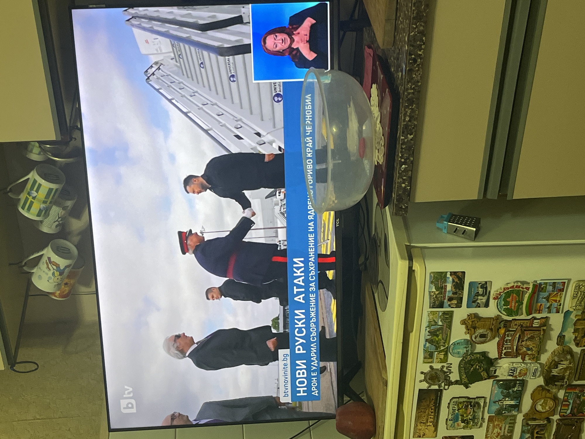 The image depicts a domestic interior, specifically a kitchen or living area, located in the city of Burgas, Bulgaria. A modern flat-screen television is centered, displaying a Bulgarian news broadcast from "bTV."
The television screen shows a main news segment featuring several individuals outdoors on an asphalt surface, possibly an airport tarmac, near a mobile staircase leading to an aircraft. One man in a dark suit with white hair and sunglasses stands prominently. A man in a dark military-style uniform with red and gold accents is seen shaking hands with a man in a dark jacket, identifiable as Ukrainian President Volodymyr Zelenskyy. In the lower portion of the screen, a blue news banner in Bulgarian reads "НОВИ РУСКИ АТАКИ" (New Russian Attacks) and "ДРОН Е УДАРИЛ СЪОРЪЖЕНИЕ ЗА СЪХРАНЕНИЕ НА ЯДРЕНО ГОРИВО КРАЙ ЧЕРНОБИЛ" (Drone hit nuclear fuel storage facility near Chernobyl). A picture-in-picture inset on the lower right displays a woman with red hair performing sign language against a blue background. The website "btvnovinite.bg" is visible on the news banner.
Below the television, on a light-colored countertop, a red apple rests on a wooden cutting board to the left. A clear, round glass bowl, possibly a fishbowl, filled with water and containing light-colored flakes, sits on a red rectangular tray with a white doily-like mat in front of the TV. Another wooden cutting board is visible to the right of the bowl.
The background reveals typical kitchen elements. Above the television, a light-colored wall or cabinet features a metal rail with hooks from which several ceramic mugs with various patterns are suspended. To the left of the television, a white refrigerator is extensively covered with numerous souvenir magnets, many depicting Bulgarian towns, landmarks, and symbols, including magnets for "ВЕЛИНГРАД" (Velingrad), "БЪЛГАРИЯ" (Bulgaria), and "Трявна" (Tryavna). A small, blue, grater-shaped magnet is attached to the side of the refrigerator. Below the countertop, light beige kitchen cabinets with horizontal grey handles are visible.